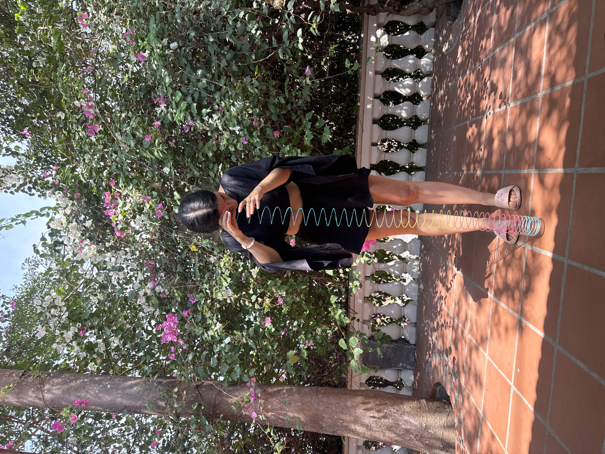 A woman is standing on a tiled terrace in Mũi Né, Vietnam. She is wearing a black outfit and pink sandals, and a colorful slinky is stretched between her head and her feet. The terrace is bordered by a white balustrade. Behind the woman is a lush bush with pink and white flowers. A tree trunk extends from the bottom of the frame, and dappled sunlight filters through the foliage onto the terracotta-colored tiles.