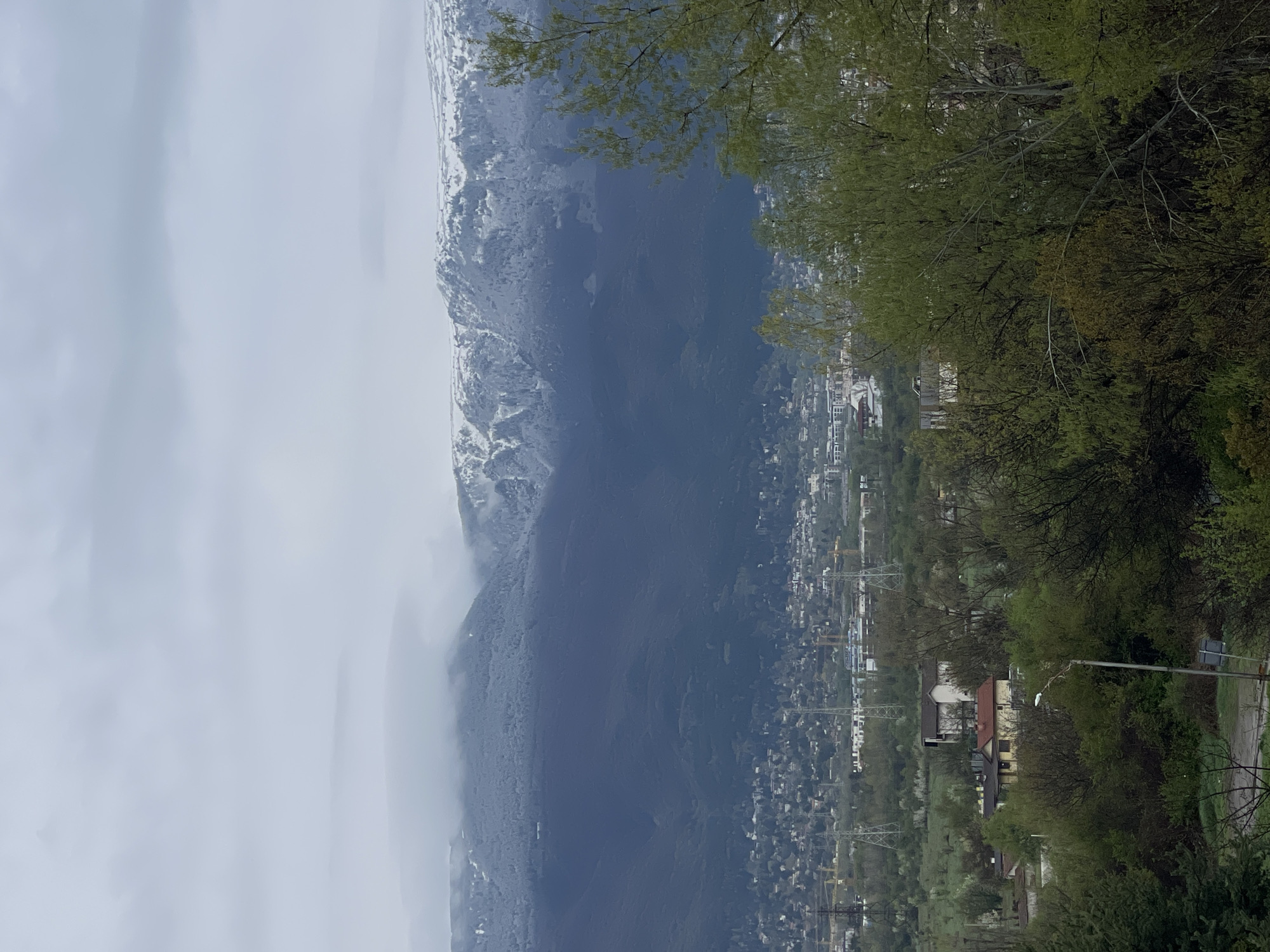 The image presents a landscape with a view of a mountain range partially obscured by clouds. Some snow is visible on the higher elevations of the mountain. In the foreground, there are trees with green foliage and urban development with buildings and infrastructure, indicating a town or city. Based on the appearance of the terrain, the urban environment, and the mountain range, it seems the location is likely in or near Sofia, Bulgaria.
