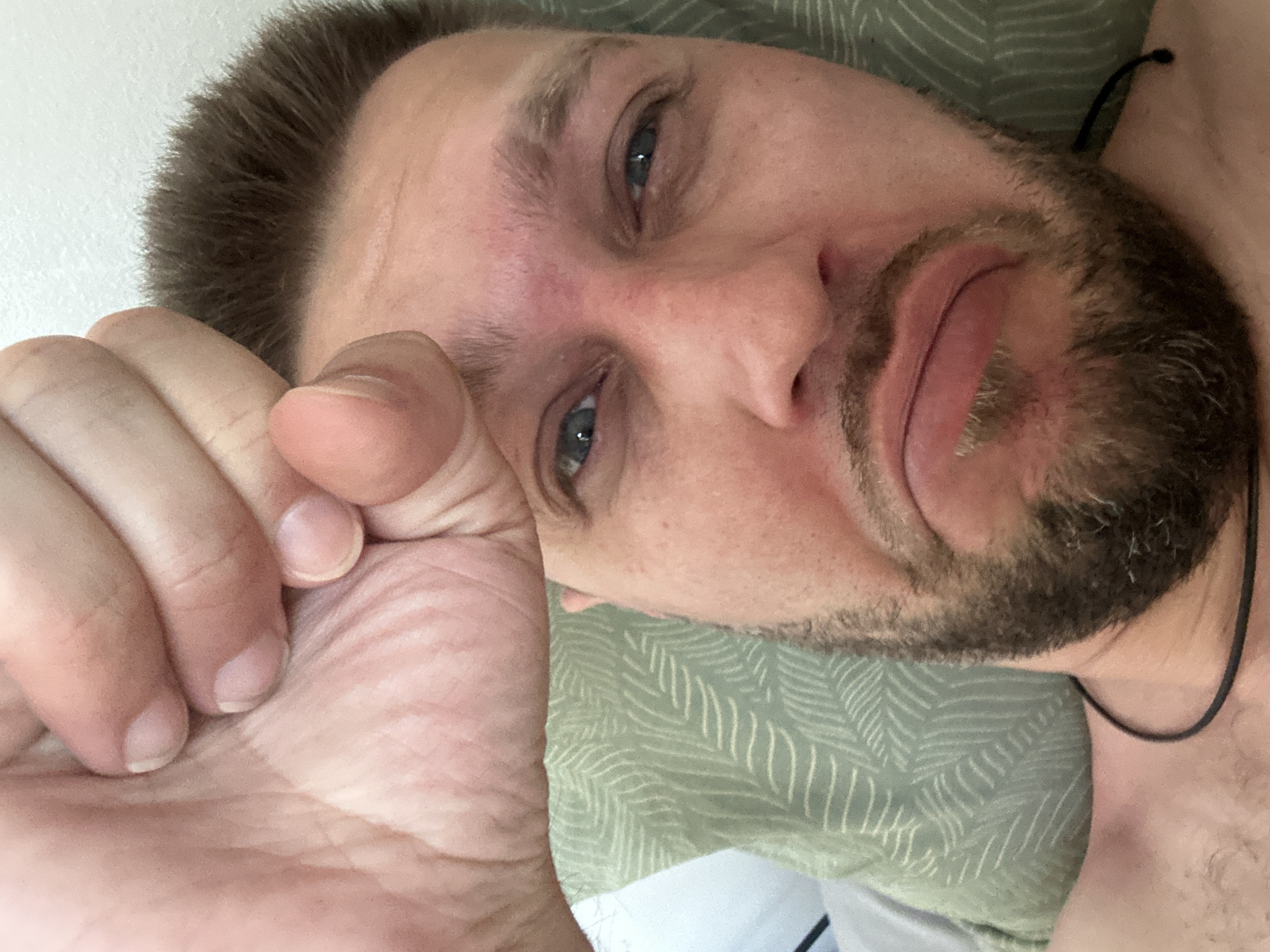 The image shows a close-up selfie of a man lying down, with his right hand covering his forehead and eyes. He has short, brown hair and a beard. His mouth is downturned, giving him a sad or pained expression. He is wearing a black cord necklace. The background consists of a green pillow with a leaf-like pattern and a white wall. There is no visible information to determine the location within Zvezdara Urban Municipality, Serbia.