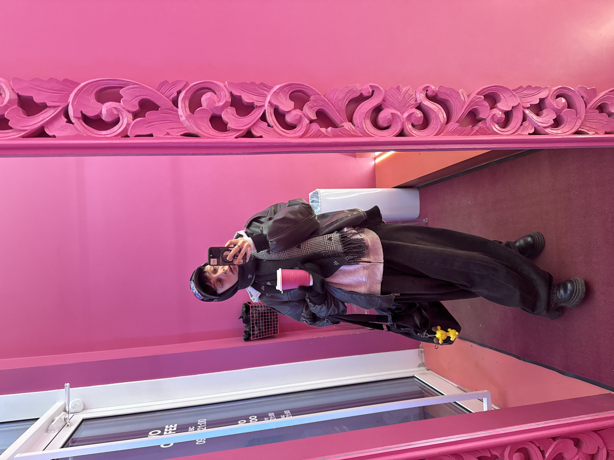 A person is taking a selfie in a pink-themed interior. The person is wearing a black cap, jacket, and baggy pants, holding a pink coffee cup in one hand and a smartphone in the other. The setting appears to be a shop or cafe in Moscow, Russia, based on the text visible through the glass door, which shows operating hours in a format common in Russia. The interior features a decorative pink frame or molding and matching pink walls. A bag and other items are also visible near the person.