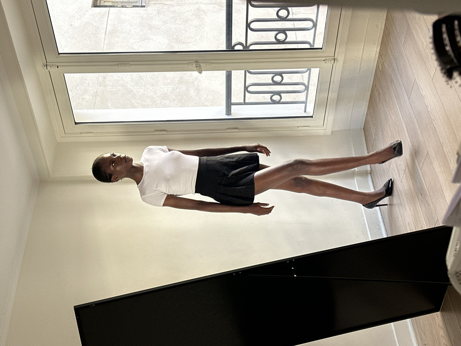 The image depicts an indoor scene within a room in Paris, France, identifiable by the distinctive Haussmannian-style double window and wrought-iron balcony railing visible on the right. The room features light-colored walls and a light-toned wooden floor. Natural light illuminates the space from the window.
A single individual, a woman with dark skin and short dark hair, stands centrally, captured in a walking or posing stance with her left leg forward. She is attired in a form-fitting white short-sleeved top, a black pleated mini-skirt, and black pointed high-heeled pumps. Her head is turned slightly to the right, with her gaze directed beyond the upper right edge of the frame.
To the left of the frame, a large, black, rectangular object, consistent with a full-length mirror or studio backdrop, stands diagonally. A portion of a dark hairbrush is visible on the floor in the bottom right corner.