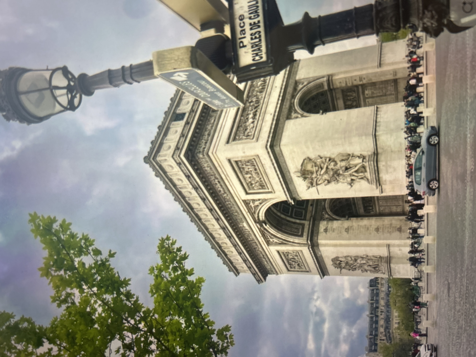 The image, taken with a vertical orientation and rotated 90 degrees counter-clockwise, captures the iconic Arc de Triomphe in Paris, France, under a cloudy sky. The monumental arch, constructed of light-colored stone, dominates the central and right portions of the frame, showcasing its classical architecture, ornate carvings, and arched passageways.
In front of the Arc, a discernible crowd of people is gathered, suggesting tourists or visitors, particularly visible queuing or walking along the right side of the monument. A light-colored car (possibly grey or silver) is parked or moving slowly on the paved street adjacent to the crowd, and a line of bicycles or scooters can be seen further back.
In the foreground, on the left side of the image, a tall, decorative black street lamp with a visible lantern fixture extends upwards. Attached to its pole are street signs. One prominent black sign clearly reads "Place CHARLES DE GAULLE". Another blue, arrow-shaped sign is partially visible below it, but its text is obscured by its angle and blur. Lush green leaves from a tree frame the bottom-left corner of the image, adding natural elements to the urban scene. Beyond the Arc de Triomphe, other buildings with typical Parisian architecture can be seen in the distance beneath the overcast sky. The scene appears to be daytime.