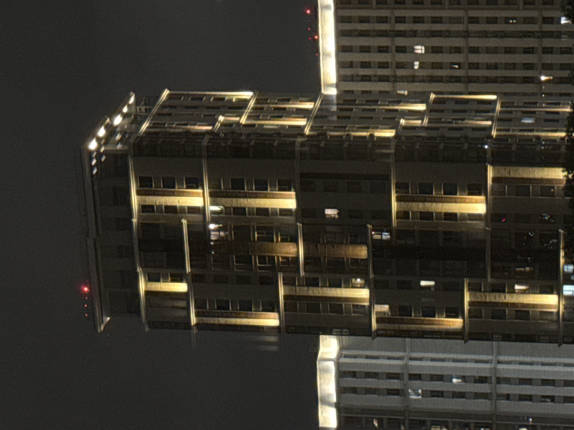 In Khimki, Russia, at night, a view shows a tall, illuminated building with a modern, rectangular design. The building is lit from within and by external lighting that highlights its vertical structure and staggered window placements. To the left, another building of similar height and style, also illuminated, is partially visible. The sky is dark and overcast, suggesting a cloudy night. The scene captures an urban nighttime landscape, focusing on the architectural features of contemporary high-rise residential or office buildings.