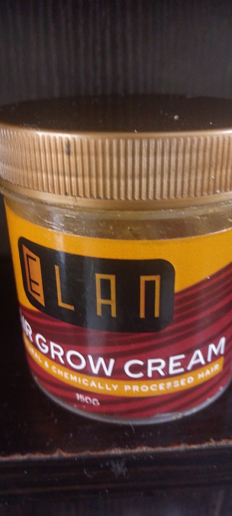 A close-up, eye-level shot shows a jar of "ELAN HAIR GROW CREAM" resting on a dark, wooden surface. The jar has a gold-colored, ribbed lid and a label that is predominantly yellow and red. The yellow section features the brand name "ELAN" in bold, orange letters against a black background. Below this, a red band with horizontal lines contains the text "HAIR GROW CREAM" in white letters. Smaller yellow text beneath that reads "NATURAL & CHEMICALLY PROCESSED HAIR". The weight, "150G", is visible at the bottom of the label. The background is dark and out of focus. There are no people or discernible city landmarks visible in this image. The lighting suggests indoor conditions, likely with artificial light sources. The image appears to have been taken in Jalingo, Nigeria.