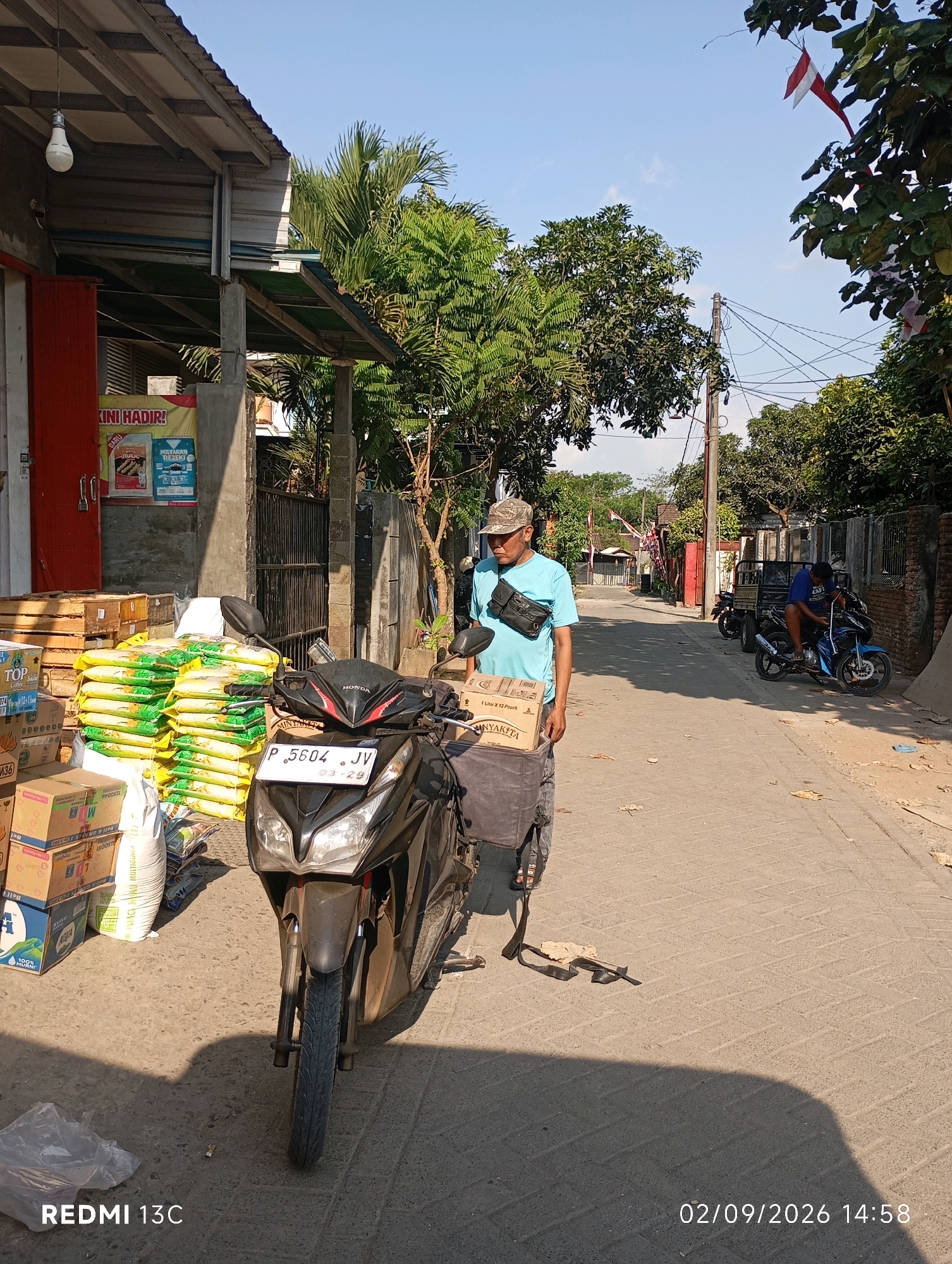 In Rambipuji, Indonesia, on a sunny day, a man wearing a light blue t-shirt and a cap stands next to a black motorcycle. He has a black fanny pack across his chest and is looking away from the camera. The motorcycle has a license plate that reads "P 5604 JV". In the background, there are stacks of yellow bags, likely containing rice or other goods, and several cardboard boxes. A building with a red door is visible on the left, and trees and power lines are present in the distance. Another man is seated on a blue motorcycle further down the street. The scene appears to be a street in a semi-urban area, possibly a market or a delivery point, given the merchandise.
