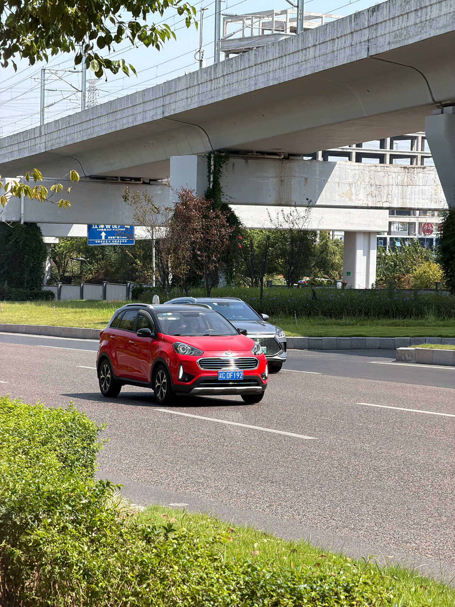 The image shows a street scene in Jiangjin, China. A red Kia SUV is driving on the right side of the road, with a silver sedan following behind it. Both vehicles have their headlights on. The road is asphalt and has white lane markings. In the background, there is a concrete overpass with power lines and vegetation growing on it. A blue sign with Chinese characters is visible on the side of the road. The scene appears to be a sunny day with clear skies.