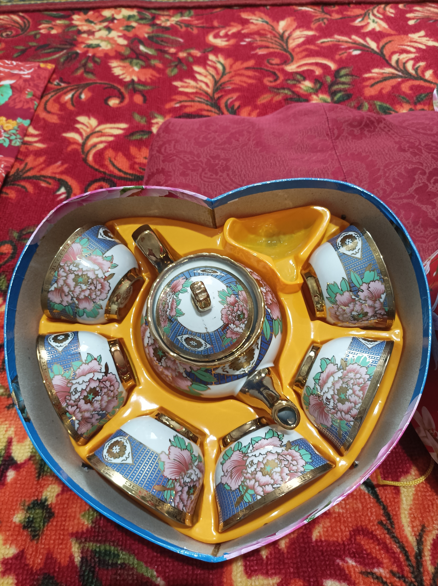 In Seýdi, Turkmenistan, a heart-shaped box containing a tea set is presented on a red, floral-patterned carpet. The tea set, with a gold-trimmed, blue, and white floral design, includes a teapot and six teacups. The teacups are arranged around the teapot within the yellow plastic insert of the box. The background is dominated by the textured red carpet and a folded red fabric.