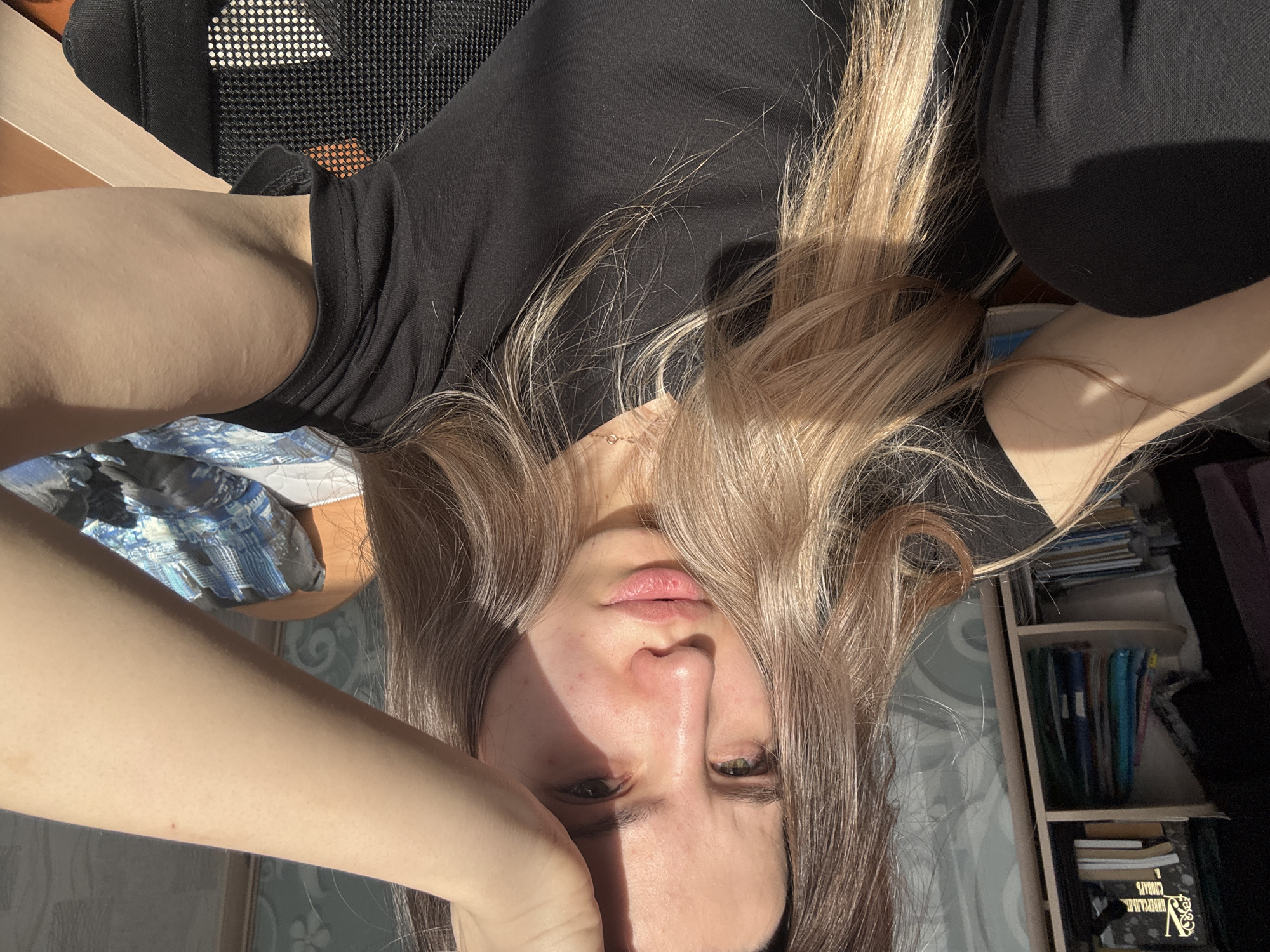 A young woman with long blonde hair is sitting in a chair. She is wearing a black top. The location appears to be an indoor space, possibly a home or office, with a bookshelf visible in the background. The city of Noviy, Russia, is not discernible from the visual information provided.