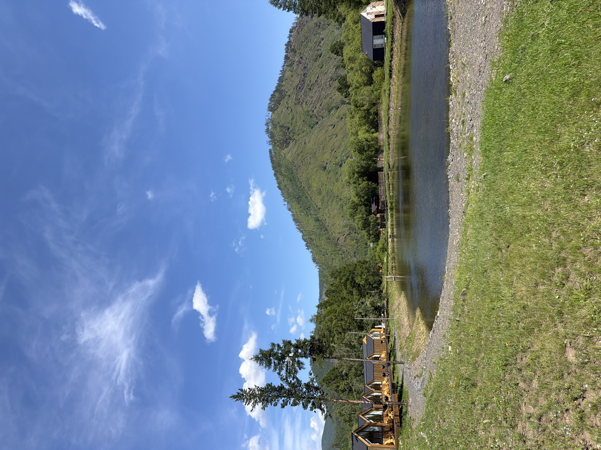 A tranquil waterscape unfolds under a bright blue sky dotted with wispy white clouds. In the foreground, a grassy meadow slopes gently towards a calm, dark body of water. The water's edge is lined with a narrow band of gravel. Along the left bank of the water, a row of modern, wooden cabins with large windows and sloped roofs are visible, each with a small wooden deck. Two tall, slender pine trees stand to the left of the cabins. Across the water, the middle ground is filled with dense green trees, and beyond them, a large, verdant mountain rises, its slopes covered in scattered trees and brush. On the far right side of the scene, near the water's edge, a small, dark-roofed building with white trim stands out against the green backdrop. A wooden fence runs along the edge of the tree line in the middle ground. The overall impression is one of serene natural beauty and peaceful retreat. The image appears to be a photograph taken in the vicinity of Bol'shoe Goloustnoe, Russia, known for its scenic landscapes along Lake Baikal.