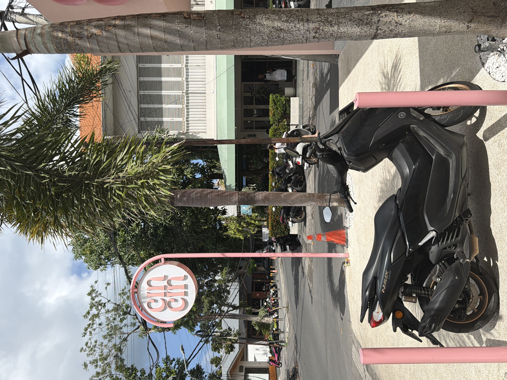 The image shows a street scene in Mengwi, Indonesia. In the foreground, a black NMAX scooter is parked on a paved area with white gravel and two pink bollards. Behind the scooter, a pink sign with the word "cin cin" in a circular design is visible, supported by a pink pole. Further back, there are several other scooters parked along the street. On the right side of the image, a portion of a pink building is visible, along with a palm tree trunk. Across the street, a building with a light-colored facade, white louvered shutters, and a green awning is present. A person wearing a white apron stands near the entrance of this building, holding a tray. Trees and power lines are visible in the background. The sky is partly cloudy.