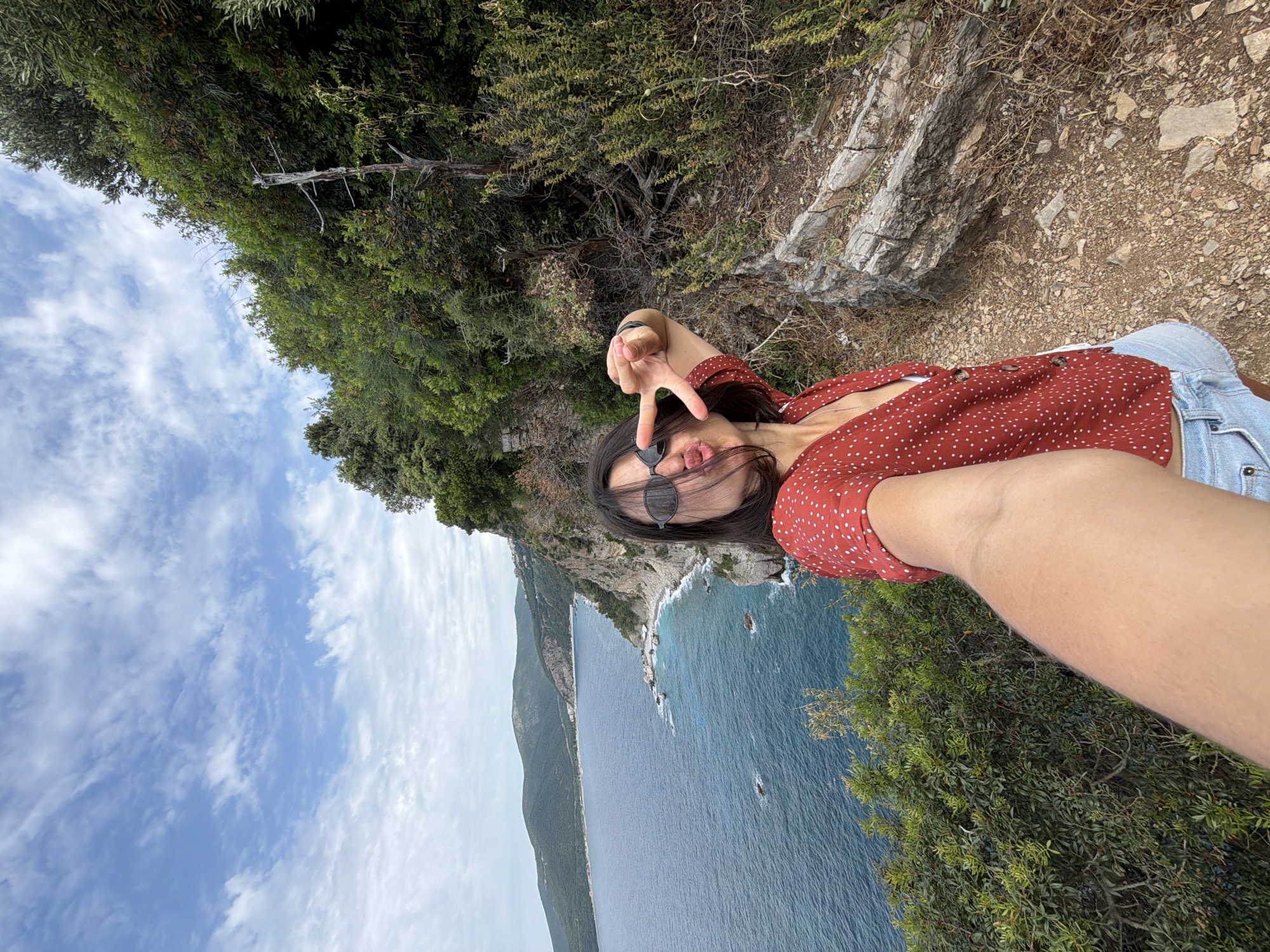 A woman captures a playful selfie amidst a breathtaking coastal landscape, likely a clifftop view in Budva, Montenegro. Positioned in the foreground, she smiles at the camera, making a peace sign with one hand and playfully sticking out her tongue. She is wearing sunglasses, a red top with white polka dots, and light-wash denim shorts.
The background reveals a stunning panorama under a bright daytime sky with scattered white clouds. To the left, a vast expanse of deep blue-green sea extends to the horizon, dotted with a few small rocks or islets. A rugged, mountainous coastline, densely covered in green vegetation, runs along the far side of the water. On the right, the immediate cliff edge is steep and covered with rich, green foliage and sturdy trees, creating a natural border to the expansive view. The ground near the woman appears rocky and earthy, suggesting a trail or lookout point. The overall scene conveys a cheerful mood and highlights the natural beauty of the coastal environment during fair weather. There is no visible text in the image.