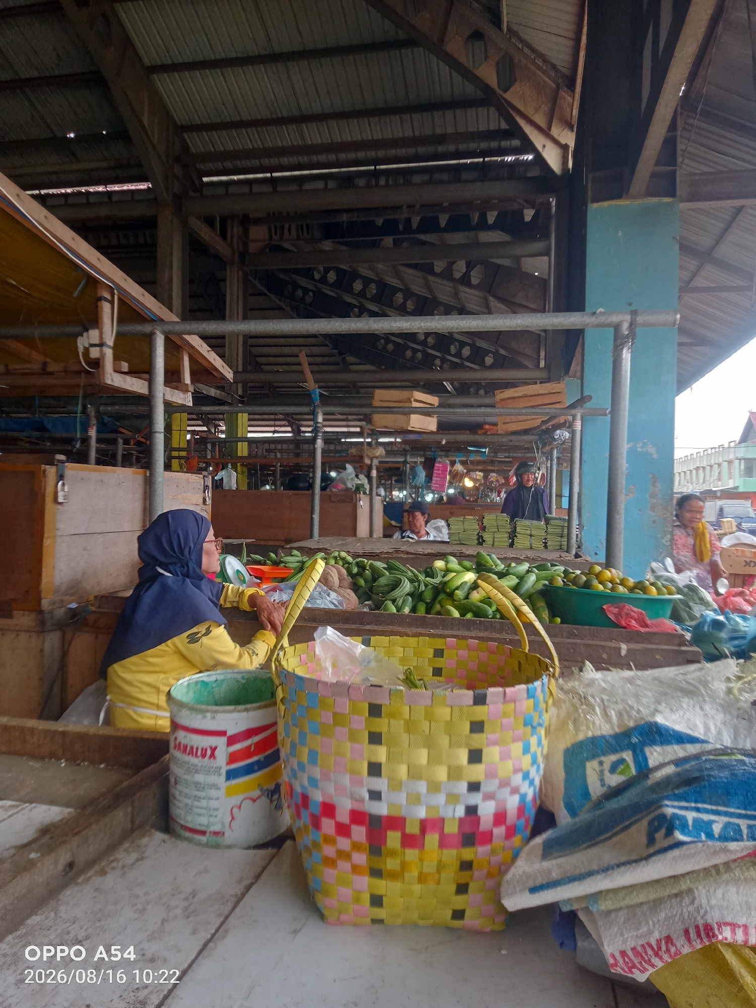 A woman wearing a dark blue hijab and a yellow long-sleeved shirt is seated at a market stall, her arms resting on the edge of a woven basket. In front of her, a white bucket with red and blue lettering is visible. The market stall displays an abundance of fresh produce, including piles of green cucumbers and a bowl of orange fruits, likely oranges or tangerines. Behind the woman, other vendors and shoppers are visible in the bustling marketplace. The market appears to be an open-air structure with a corrugated metal roof supported by wooden beams. The scene suggests a typical day in a vibrant local market in Jambi, Indonesia, where vendors are engaged in selling fresh goods to customers.