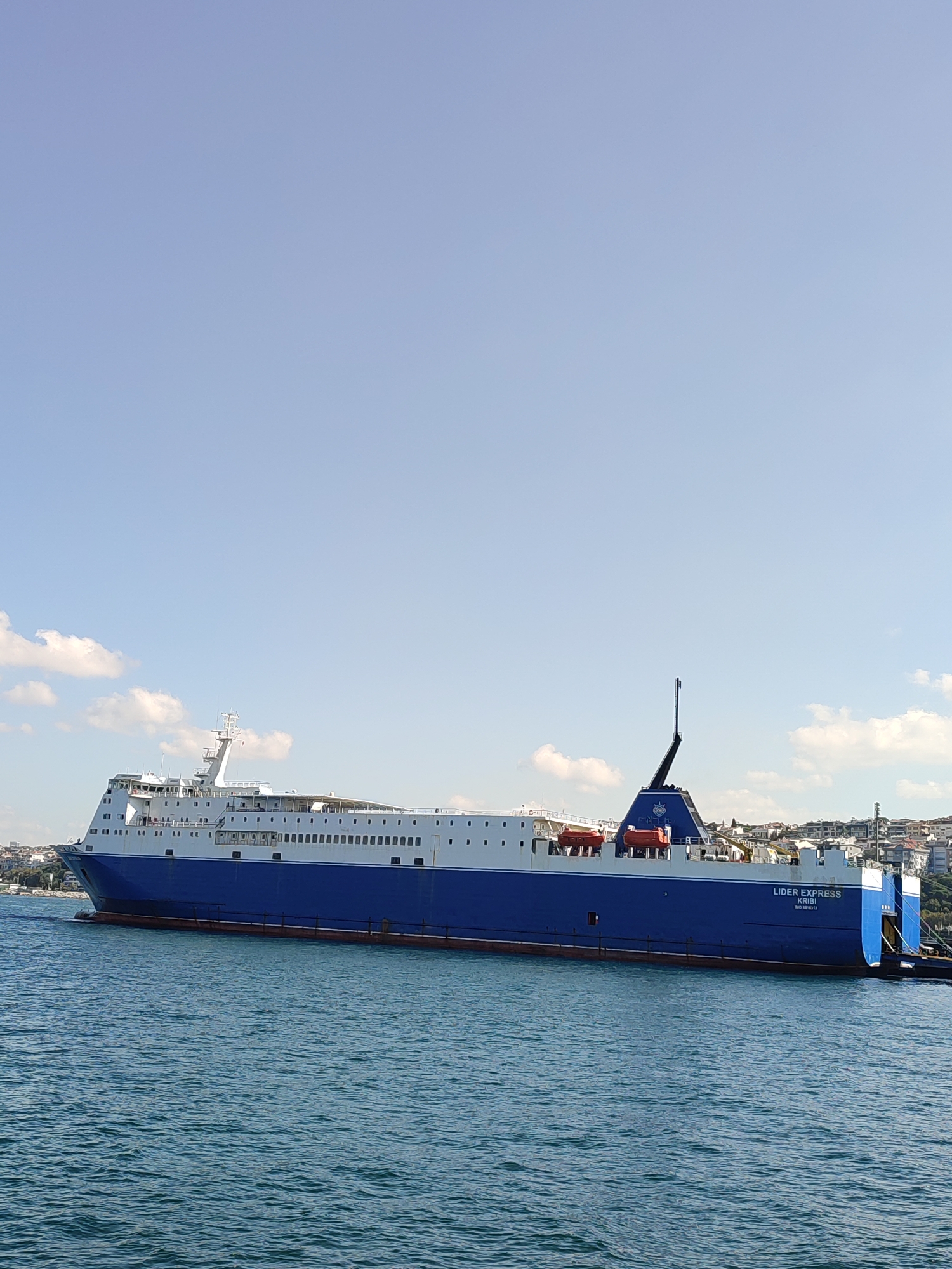 A large blue and white ferry named "LIDER EXPRESS KRIBI" is docked in the waters of Üsküdar, Turkey. The ferry is positioned to the right of the frame, with its bow extending out of view to the left. The vessel has multiple decks and windows, with life preservers visible on the upper levels. The stern of the ferry is dark blue and features the vessel's name and IMO number. In the background, the coastline of Üsküdar is visible, with buildings and trees dotting the hillside under a clear blue sky with scattered white clouds. The water is calm and reflects the blue of the sky and the ferry.