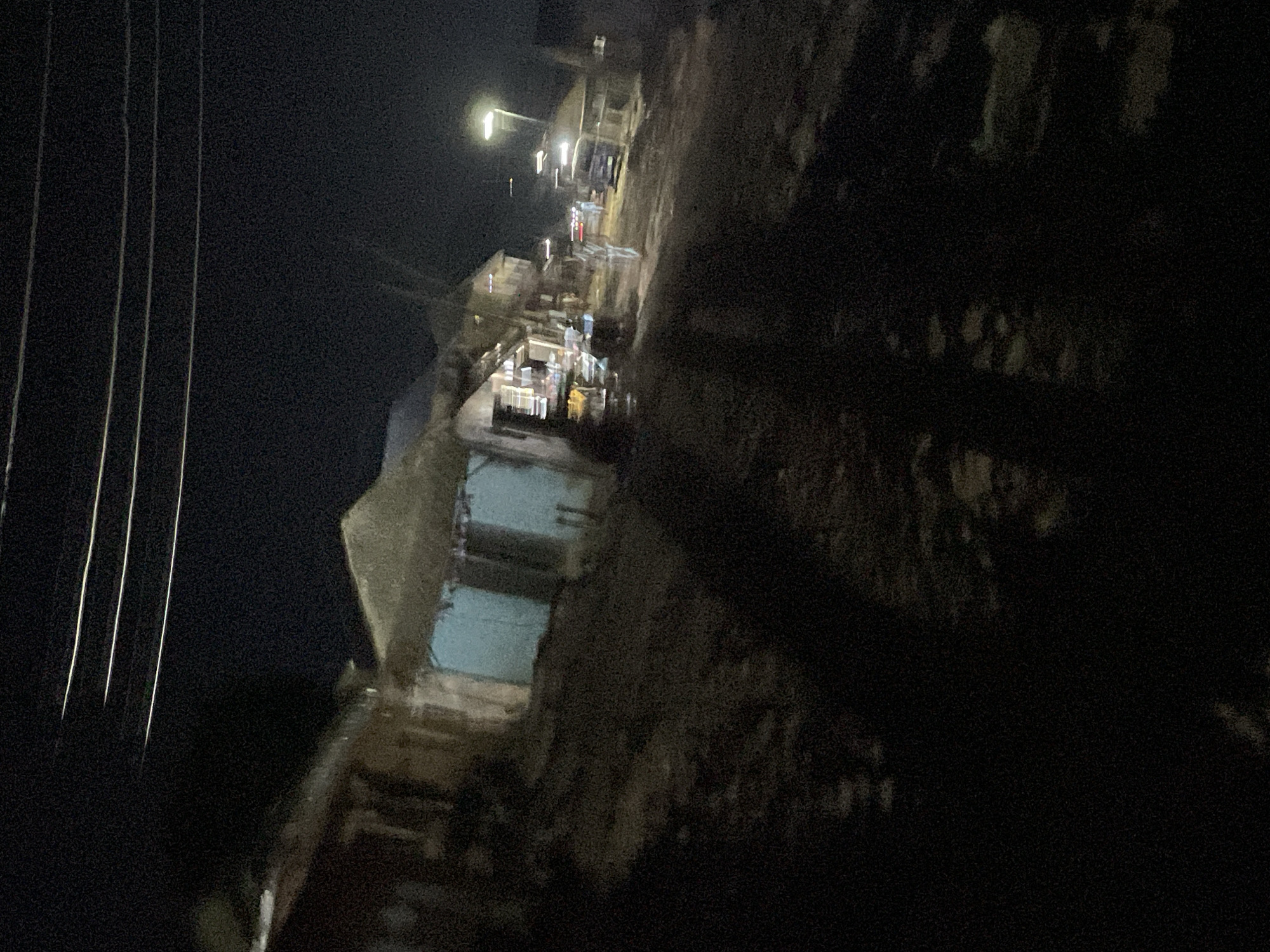 The image depicts a nighttime scene on a dirt road in Lokoja, Nigeria. The road is lined with informal structures, likely market stalls or small shops, with corrugated iron roofs and some appearing to have thatch. The buildings are dimly lit from within, casting a faint glow. Several vehicles, including what appears to be a van and other cars, are visible on the road, their headlights and taillights creating streaks of light due to the long exposure or motion. A streetlamp illuminates a portion of the scene. Shadows are cast across the road, obscuring details of the ground and any pedestrians. The overall impression is of a bustling, albeit low-light, commercial area operating after dark.