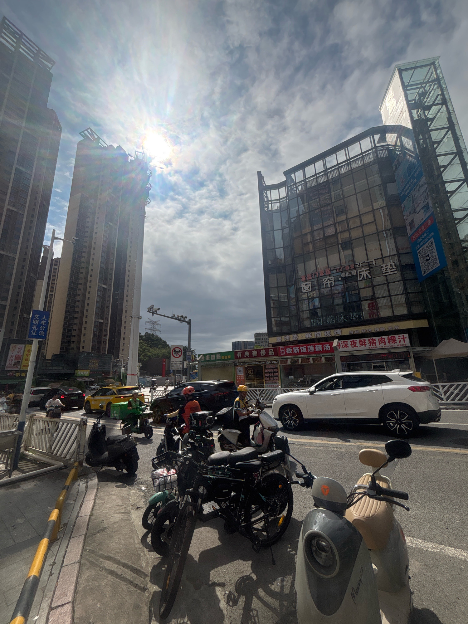 The image depicts a street scene in Nanhai District, China, characterized by high-rise buildings, moderate traffic, and numerous parked and moving scooters and motorcycles. The sun is bright, casting lens flares and highlighting the cloudy sky. Tall, modern buildings dominate the background, with one featuring a glass facade. In the foreground, the sidewalk is lined with parked electric scooters and bicycles. The street itself has several vehicles, including a white SUV, a yellow taxi, and multiple scooters. People are visible on some of the scooters, some appearing to be delivery personnel due to their bright green uniforms. One scooter has a rider wearing a yellow helmet, and another has a rider in an orange helmet. A blue sign with Chinese characters is visible on a pole on the left side of the image. The overall impression is a busy urban environment with a prevalence of two-wheeled vehicles.