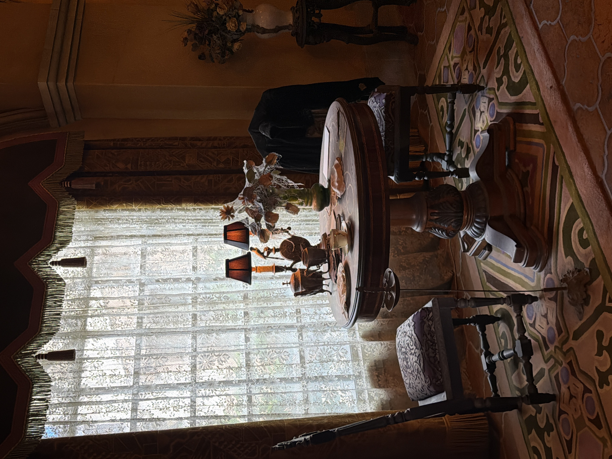 The image shows an indoor scene with ornate furnishings. In the foreground, a circular wooden table with intricate carvings is visible, topped with a multi-armed lamp featuring dark shades and decorative elements. Beside the table, a wooden chair with a patterned cushion is positioned. Behind the table and chair, a large window is covered by sheer lace curtains that let in diffused light, suggesting it is daytime.
To the left of the window, a curtain with tassels hangs down. The room's walls are decorated with a tapestry or patterned wallpaper in earthy tones and geometric designs. Above the window, architectural details are visible, including what appears to be a molded ceiling or archway. On the far left of the image, a dark cloth is draped over something, possibly furniture or a person, though no individuals are clearly depicted.
In the background, on a shelf or surface, a white vase with dried flowers is partially visible. The overall impression is one of an antique or elaborately decorated interior, perhaps a dining room or lounge. The lighting is subdued, creating a warm and somewhat dramatic ambiance. No text is visible in the image. The location context of Chessy, France, might suggest a historic building, a hotel, or a residential interior with European influences.
