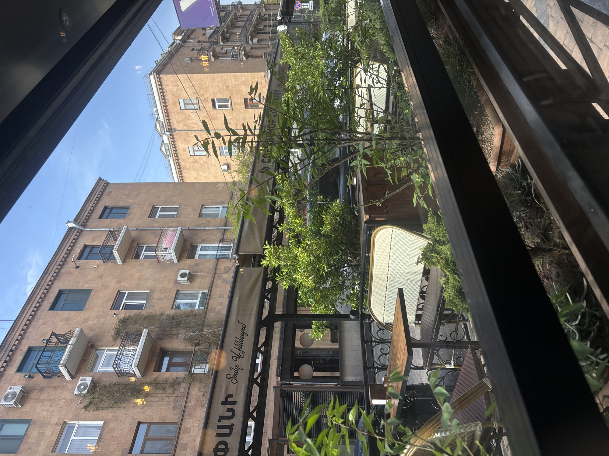 View from inside a building, looking out onto an outdoor cafe area in Yerevan, Armenia. Adjacent brick buildings with balconies are visible. The cafe has metal framework with plants and dining furniture. The sky is visible above the buildings.