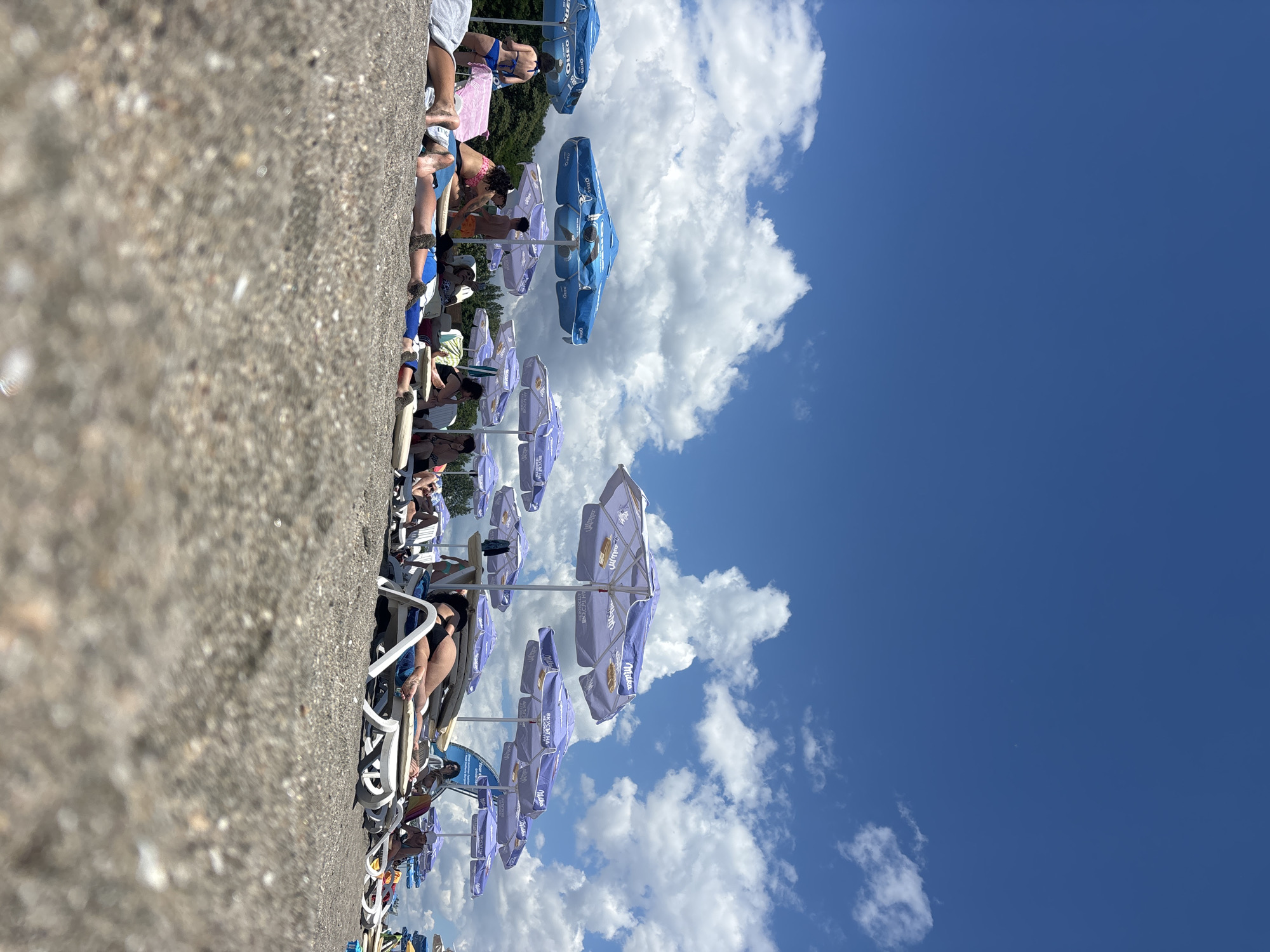 The image captures a crowded beach scene under a bright blue sky dotted with fluffy white clouds. Numerous beach umbrellas, primarily in shades of blue and purple, provide shade for beachgoers lounging on sunbeds. People are seen in various states of relaxation, some lying down, others sitting. The foreground is dominated by the sandy beach. In the background, a line of trees can be observed. The overall impression is one of a popular beach destination on a sunny day.