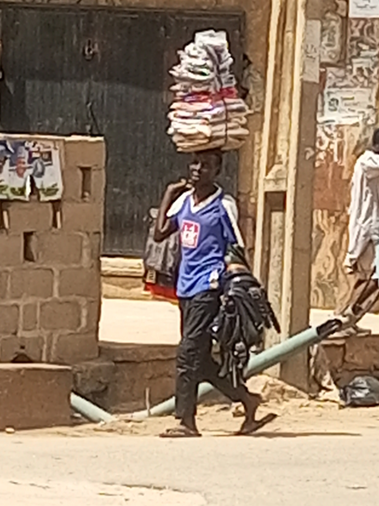 A mid-shot photograph captures a young man walking along a dusty path in Maiburiji, Nigeria, under bright daylight. He is positioned slightly left of center, moving towards the right side of the frame. The man has dark skin and is wearing a blue and white short-sleeved jersey-style top with a red and white logo (possibly "2GO" or similar) on the chest, black trousers, and flip-flops. He carries a large, irregularly shaped bundle, appearing to be stacked papers, cardboard, or fabric tied with rope, balanced on his head. A dark, possibly bag-like object is also slung over his left shoulder or arm.
The setting consists of rough-textured, light brown brick or mud-brick walls on both sides. The wall on the left has several posters or papers affixed to it. In the center-back, a dark, double-doored entrance is visible. The wall on the right displays faded patterns or old posters. Another person, partially visible in light-colored clothing, walks in the distant right background. The ground is a light brown, dusty surface, with a light green/grey pipe running horizontally in the mid-ground. Strong shadows suggest a clear, sunny day. The man appears to be engaged in transporting goods, a common activity in such settings. No specific text is clearly legible beyond the general shape of the logo on his shirt and the vague shapes on the posters.