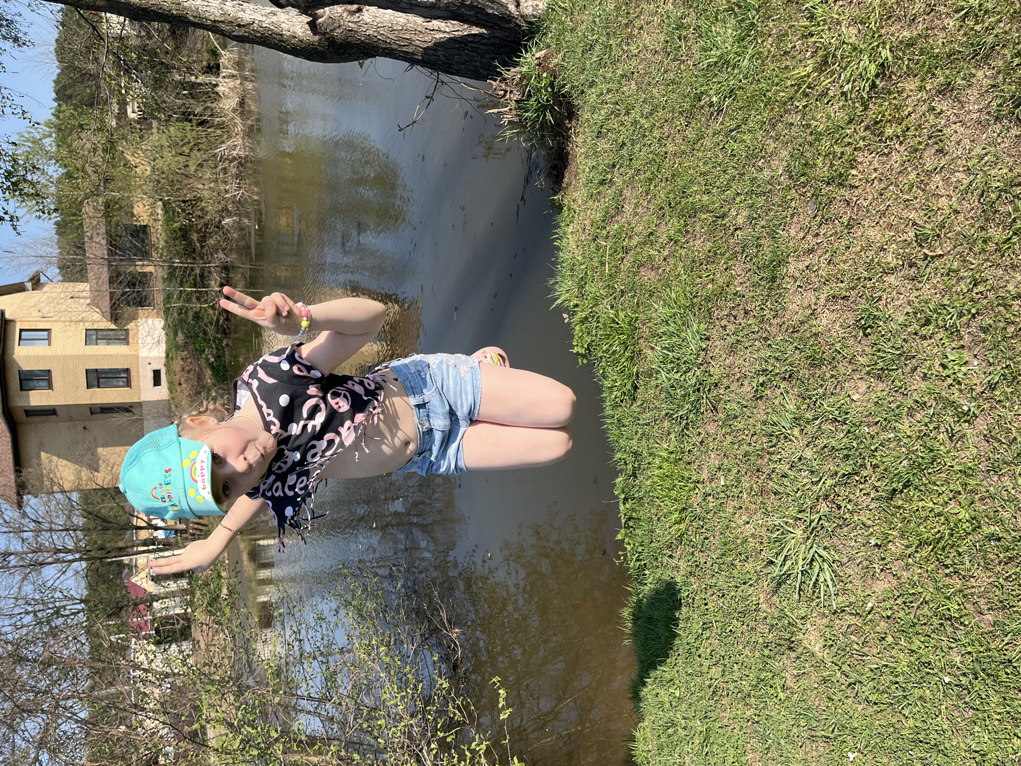 A girl in a blue baseball cap, a black-and-pink top, and denim shorts is positioned near a body of water. She is making a peace sign with both hands. The background includes a building, trees, and grass. The location is Chaikovsky, Russia.