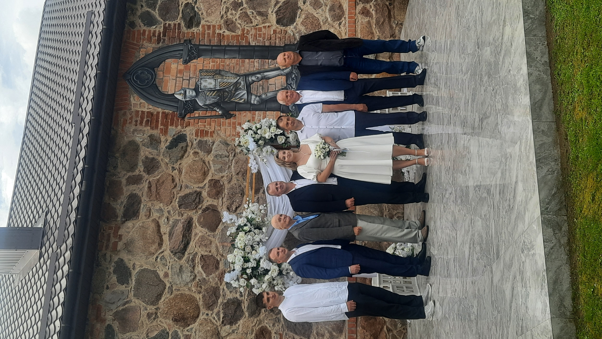 A wedding party of nine people stands on a tiered, marble-like platform in front of a stone and brick wall with a mural of a knight. The individuals are positioned from left to right: a young man in a white button-down shirt and black pants; a man in a dark blue suit; an older man in a gray suit and blue shirt; a man in a black suit; the bride in a white dress with puffy sleeves and holding a small bouquet of white flowers; a man in a white button-down shirt and dark pants; a man in a dark blue suit and dark pants; and a man in a black jacket and dark jeans. The bride is standing in the center of the group. The backdrop features floral arrangements of white roses and greenery. The mural of the knight is framed by an arched gothic structure. The environment appears to be outdoors, with a glimpse of green grass in the foreground. The location is identified as Жодзішкаўскі сельскі Савет, Belarus.