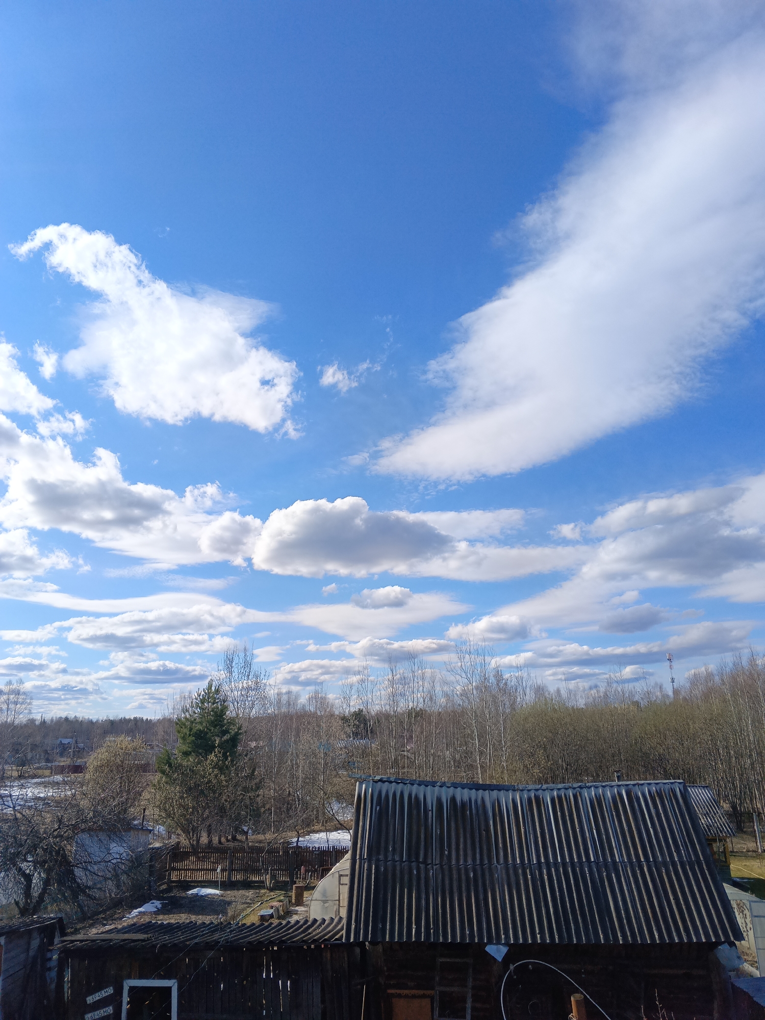 The image shows a rural scene in Красный Маяк, Russia, on a clear day with a blue sky scattered with white cumulus clouds. In the foreground, there is a wooden building with a dark, corrugated roof and a visible window. Behind the building, there are trees with bare branches, suggesting it is early spring. A partially snow-covered area is visible, possibly a body of water or field. In the distance, a telecommunications tower is visible among the trees. The overall impression is a quiet, rural landscape.