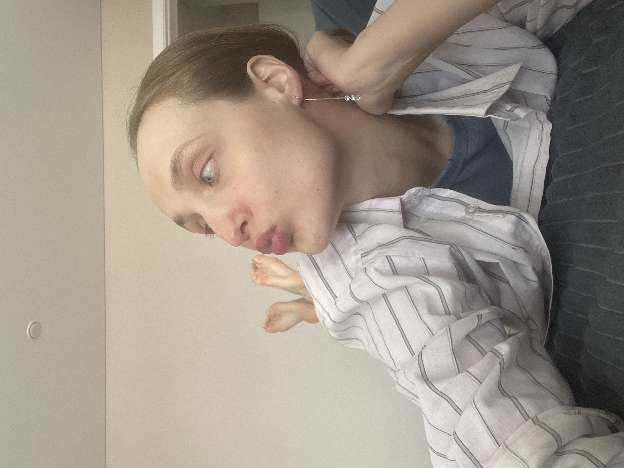 A woman is shown from the chest up, lying sideways. She wears a light blue undershirt beneath a white pinstripe shirt. A hand is inserting a long, thin earring with three crystals into her left ear. She is lying on a dark grey striped cushion or sofa. The background is a light beige wall. The location, Sepych, Russia, is extraneous information as it cannot be determined from the image.