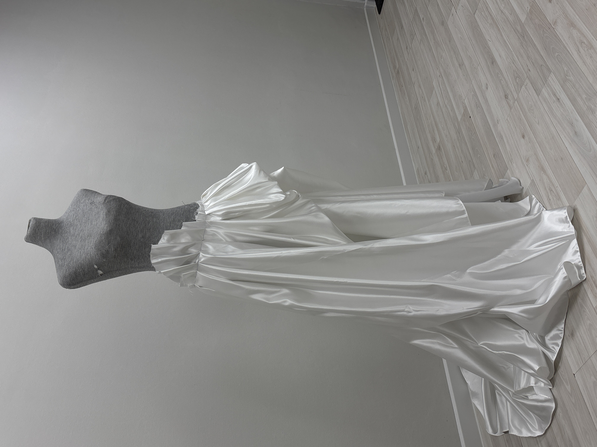 A gray dressmaker's form is draped with a long, white satin gown. The gown is gathered at the waist and flows out into a train that rests on a light wood-grain laminate floor. The scene is set against a plain, light gray wall. There is no evidence indicating the location of the image is in Perm, Russia.
