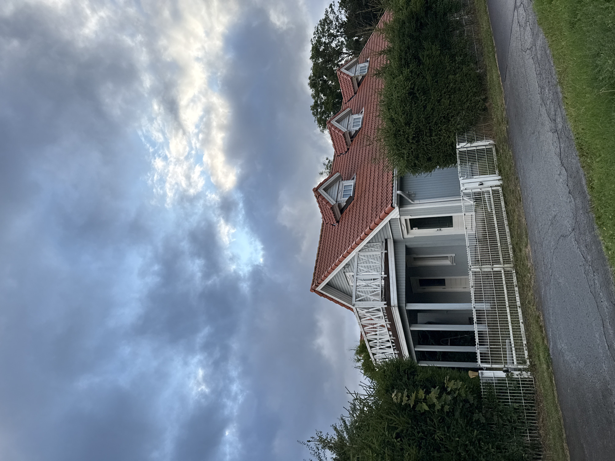 The image depicts a light blue, two-story house with a red tile roof, situated in the rural setting of Peetri alevik, Estonia. The house features a prominent white balcony with decorative railing on the upper level, supported by blue columns. Several dormer windows are visible on the roof. The property is enclosed by a white metal fence, and dense green hedges line the front and sides of the house. A paved road runs in front of the fence, with a strip of green grass to the right. The sky is overcast with dramatic clouds, suggesting a potentially stormy or dusk-like atmosphere. No people are present in the image.