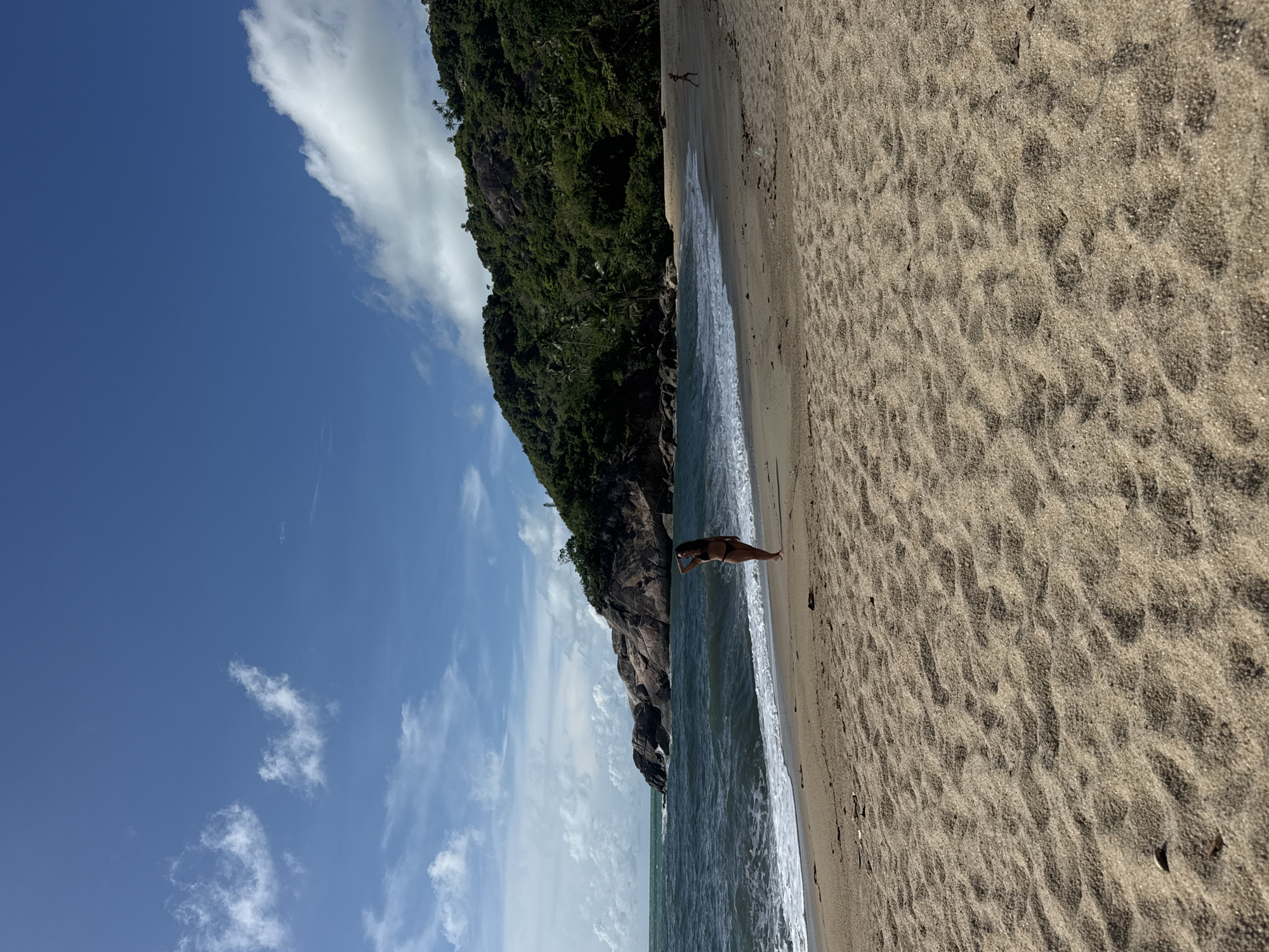 A horizontal outdoor shot shows a beach, the ocean, and a rocky, vegetated headland in Baan Thong Nang, Thailand, under a blue sky with scattered clouds. The sandy beach takes up the right half of the image, and the ocean, with visible waves, runs along the center. A single individual stands on the beach, closer to the ocean's edge. The rocky headland, covered in dense green vegetation, rises from the left side of the ocean. The sky, with scattered white clouds, fills the top left quadrant of the image.