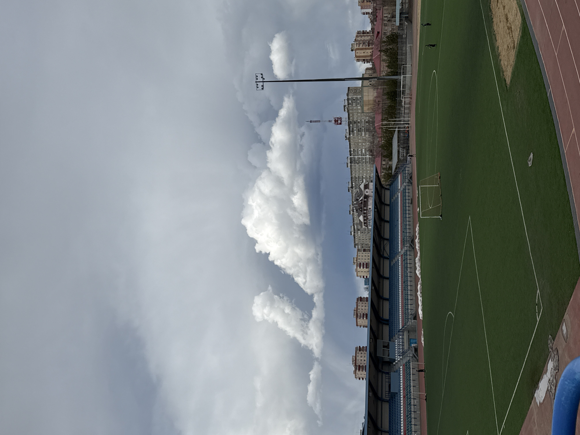 The image shows a soccer stadium in Mirny, Russia, on an overcast day. The sky is dominated by gray clouds, with a few patches of blue visible. A large bank of cumulonimbus clouds sits near the edge of the city. A sports complex is situated adjacent to residential buildings. On the right of the frame, an artificial turf soccer field is visible, marked with white lines. Adjacent to the field is a red running track. Empty blue seats are in the stadium's stands. Tall light posts stand on the city side of the stadium. Several buildings are on the horizon. Three people are walking near the field.