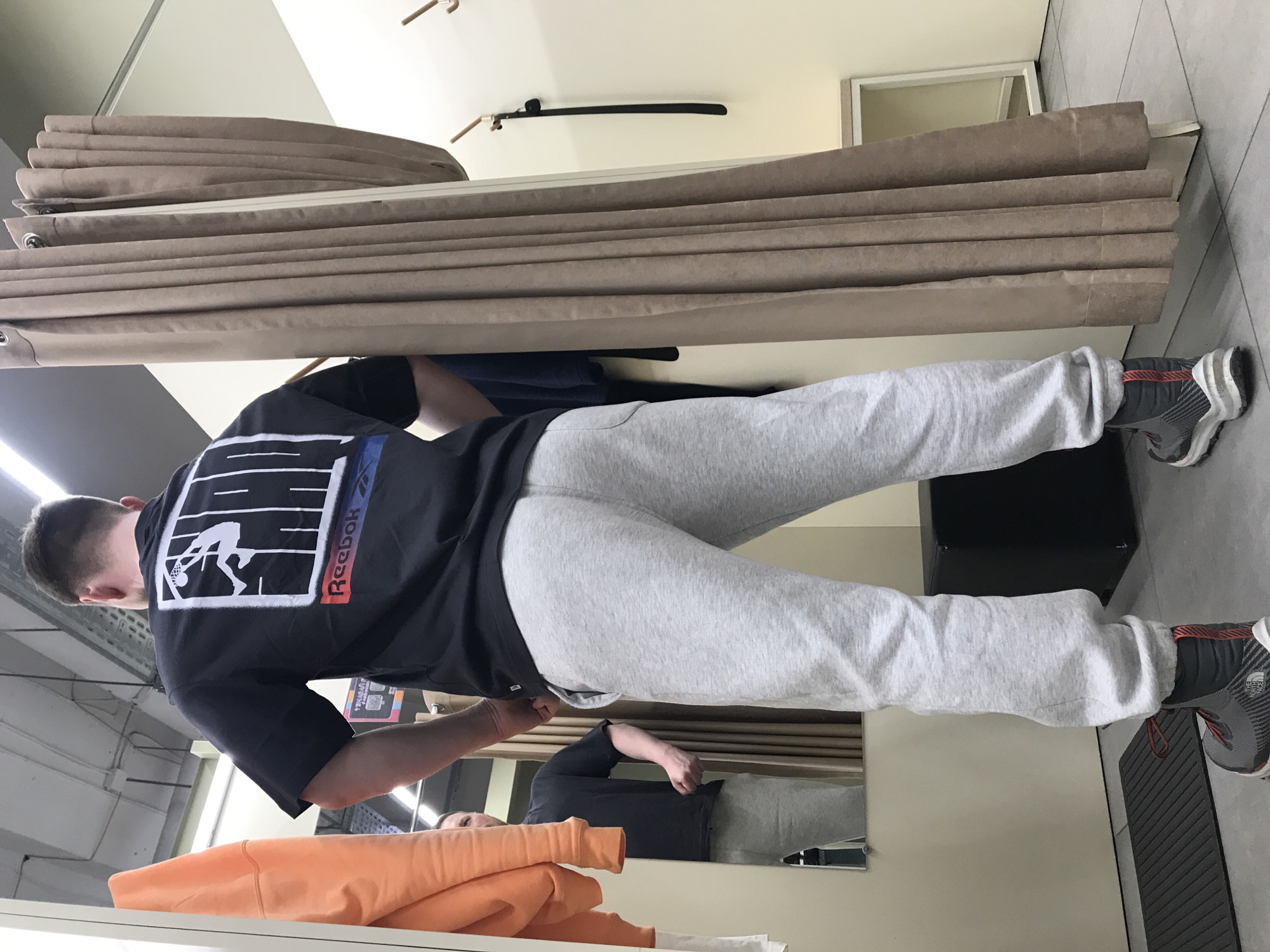 A man stands inside a dressing room. He wears gray sweatpants, a black t-shirt with a "Reebok" logo, and gray sneakers. The dressing room has beige curtains. Other clothing items are visible, including an orange hoodie. The location appears to be in Perm, Russia, though this is not directly verifiable from the image.