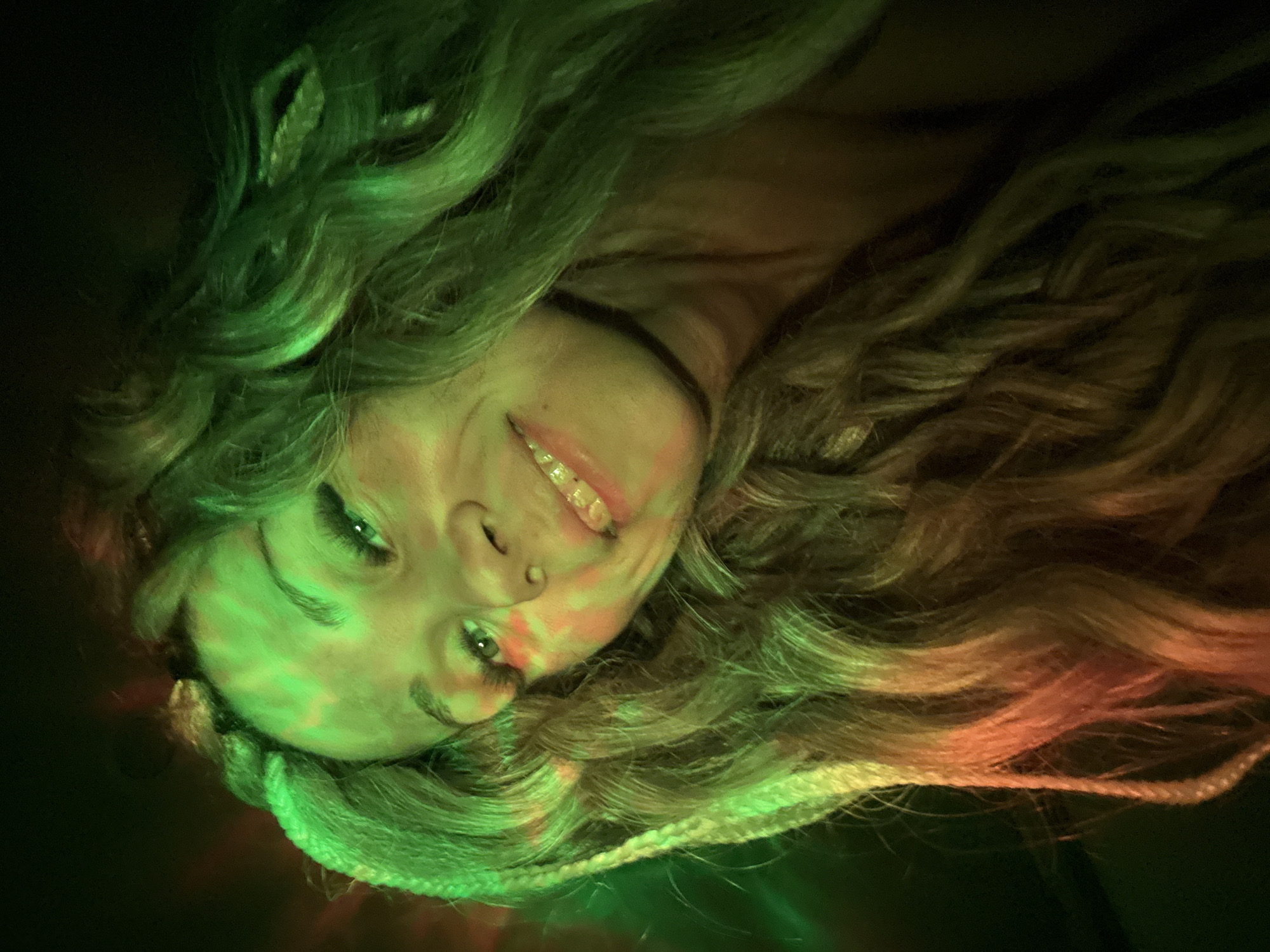 A woman lies on her back, illuminated by green and red light. Her blonde hair is styled in wavy sections and braids. The location is indiscernible.