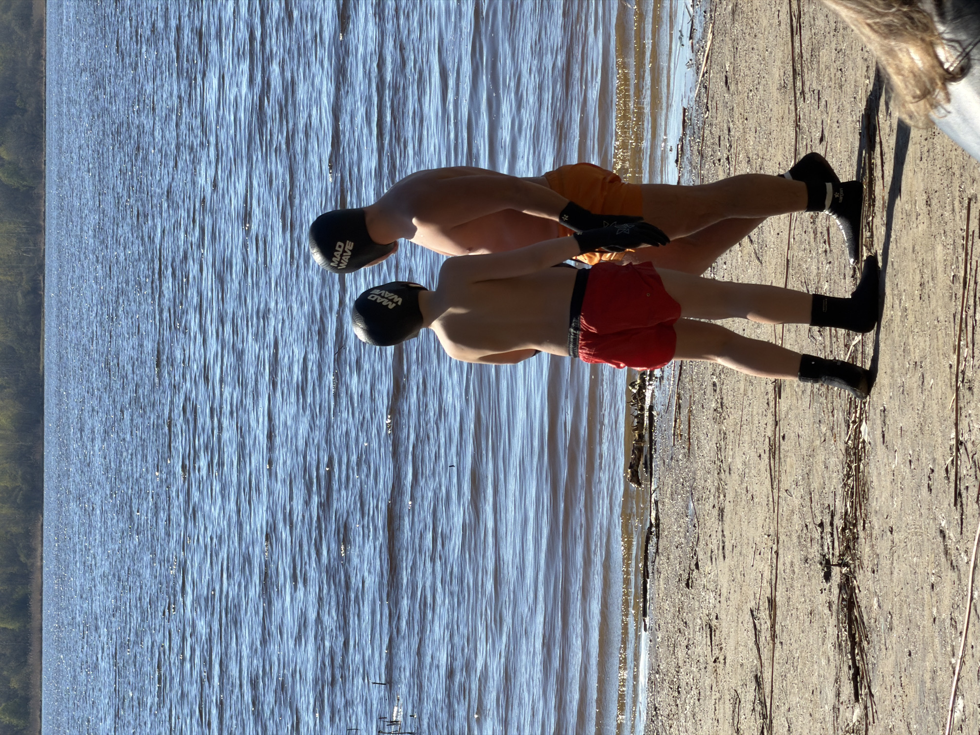 Two shirtless individuals in swim caps and swimming gear stand on a sandy beach next to a body of water. The person in the foreground wears red swim trunks, while the person behind them wears orange swim trunks. Both are wearing black swim caps with white lettering and black socks and water shoes. The water is a dark blue with ripples and reflections of sunlight. In the background, there is a visible shoreline with trees. The location is likely Saint Petersburg, Russia due to the presence of a sandy beach and a large body of water with trees in the background.