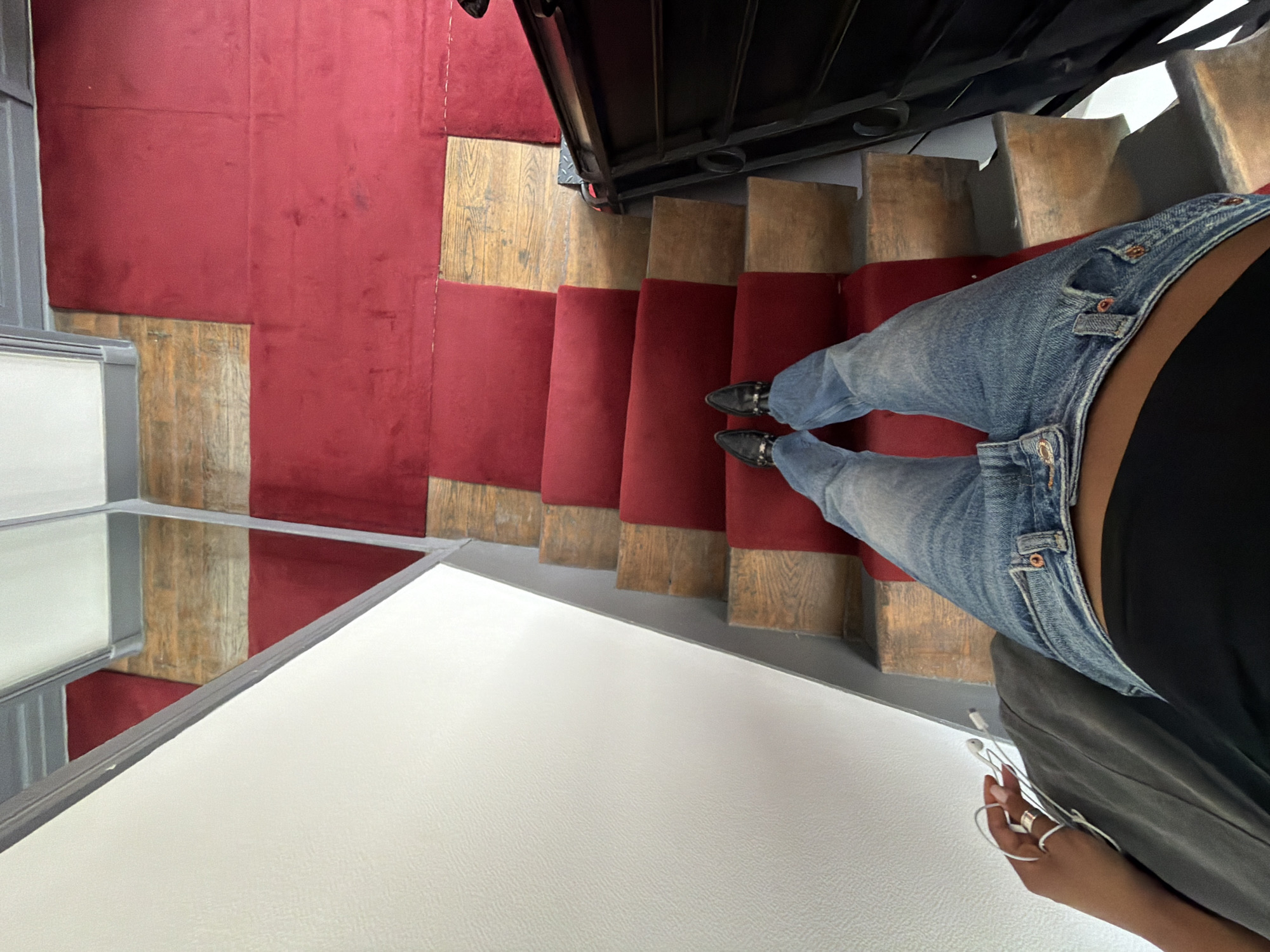 The image shows a person's legs and feet standing on a set of stairs. The stairs have wooden treads with red carpet runners. A white wall is visible to the left, and a black railing is to the right. The person is wearing blue jeans and black pointed-toe shoes. They are holding white earbuds in their left hand. The scene appears to be indoors, possibly in a residential building or a shop. There is no visual information in the image that indicates the location is Paris, France.