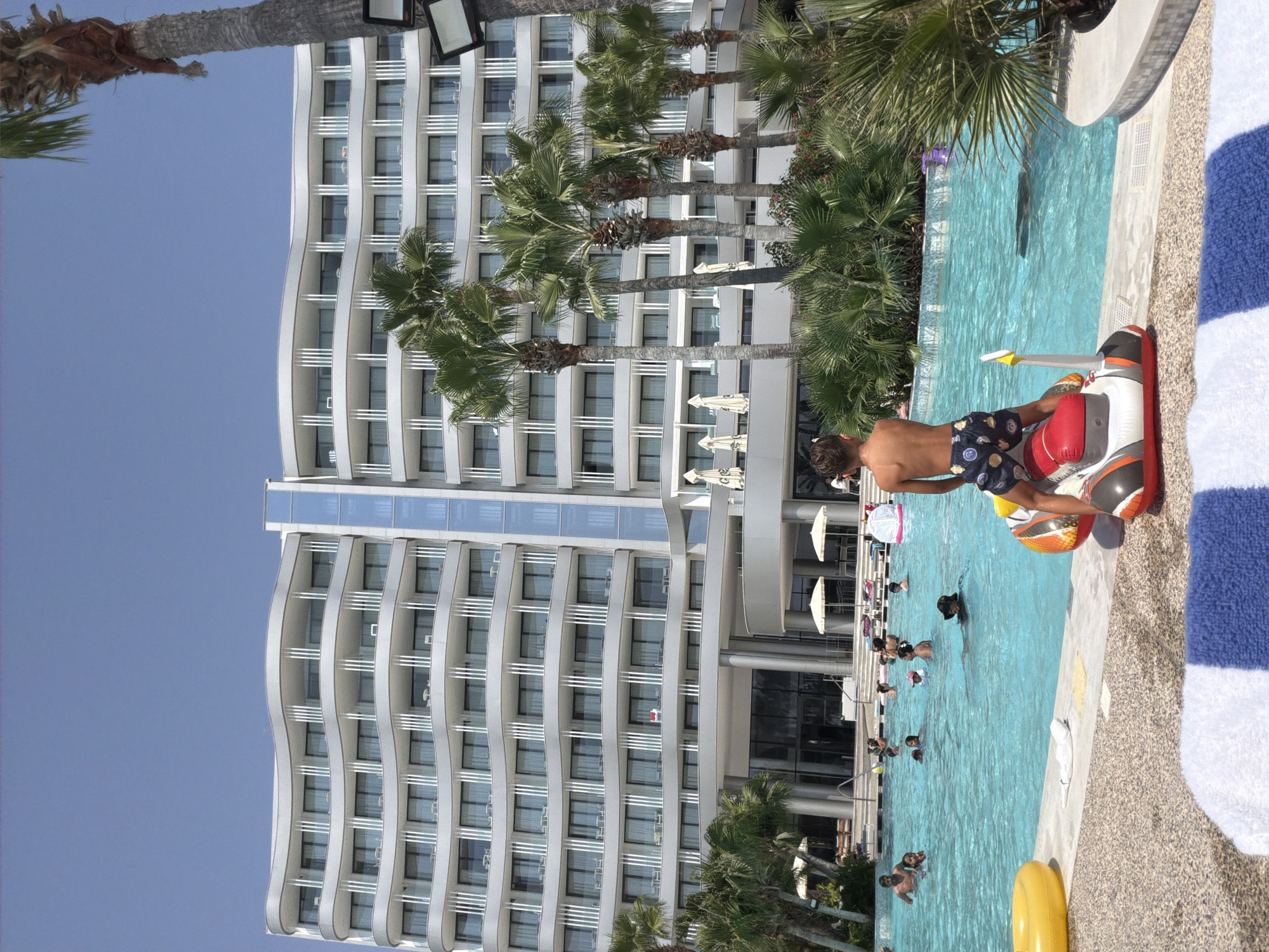 The image depicts a lively scene at a resort swimming pool, with a large, modern hotel building in the background. The hotel features a distinctive wave-like architectural design with multiple balconies and windows. Palm trees are scattered around the pool area, and the sky is clear and blue.
In the foreground, a young boy stands on an inflatable toy shaped like a boat. He is wearing patterned swimming shorts and facing away from the camera, looking out at the pool. To his left, a yellow inflatable ring floats in the pool.
The swimming pool itself is filled with people of various ages, some swimming, others wading, and some interacting in the water. The water is a bright turquoise color. Scattered throughout the pool are various inflatable toys, including a large circular one in the background. Several lounge chairs and umbrellas are visible on the pool deck.
The overall atmosphere is one of recreation and relaxation on a sunny day. The resort appears to be located in Oroklini, Cyprus, a popular tourist destination known for its coastal resorts.