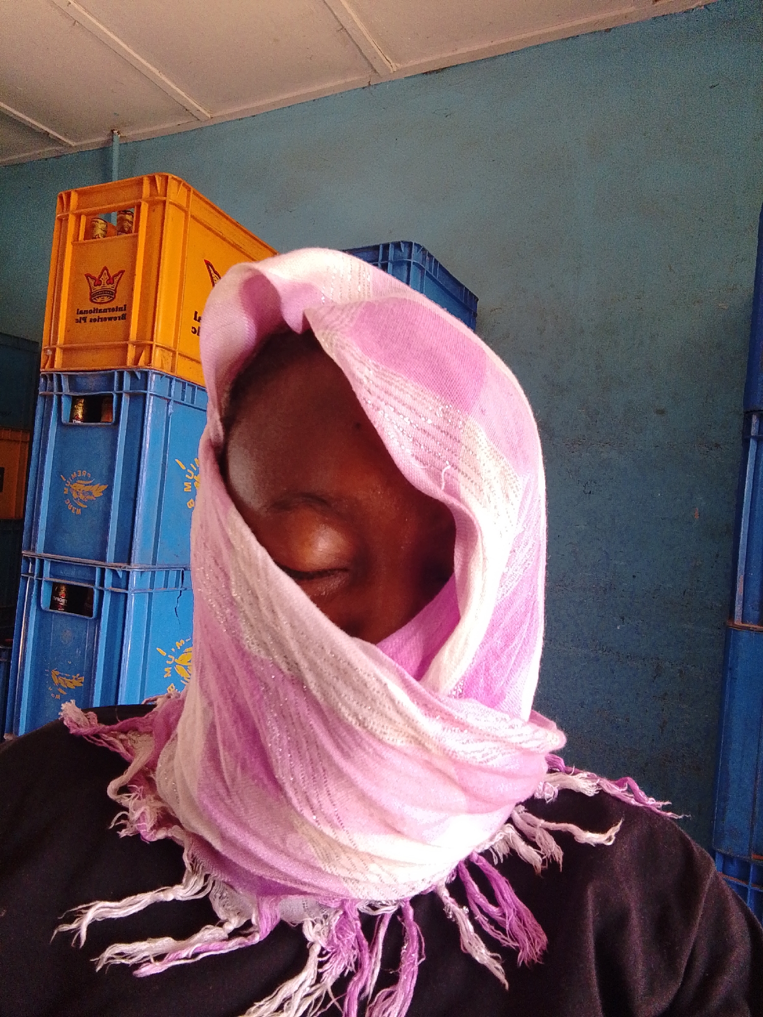 A person wearing a pink and white headscarf stands in front of stacked yellow and blue beverage crates. The person's face is mostly covered by the headscarf, with only the closed eyes visible. The background consists of a light blue wall and blue beverage crates. The location is Unknown, Unknown.