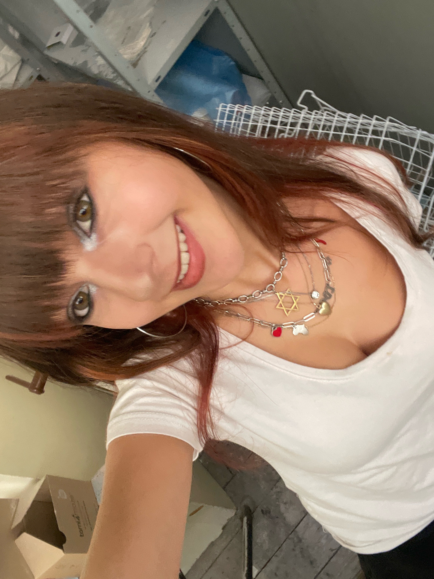 The image shows a selfie of a woman with long, reddish-brown hair and bangs, wearing a white t-shirt and a multi-layered silver necklace with various charms, including a Star of David and heart shapes. She has on makeup, including eyeshadow and lipstick, and is smiling at the camera. The background appears to be an indoor space with shelving units, some boxes, and a wire rack. The lighting suggests it might be an unfinished or storage area. There is no information about the location within the city of Raanana, Israel, or any specific details about the environment that would indicate such a location.