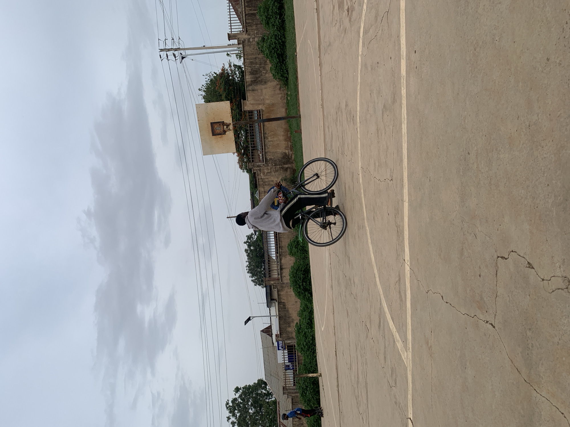 The image depicts an individual riding a bicycle on a cracked concrete basketball court in Jos, Nigeria. The person is wearing a light-colored sweatshirt and black pants. A weathered basketball hoop stands at the edge of the court, adjacent to a concrete building and vegetation. Overhead, power lines stretch across a cloudy sky. In the background, additional buildings and trees are visible.