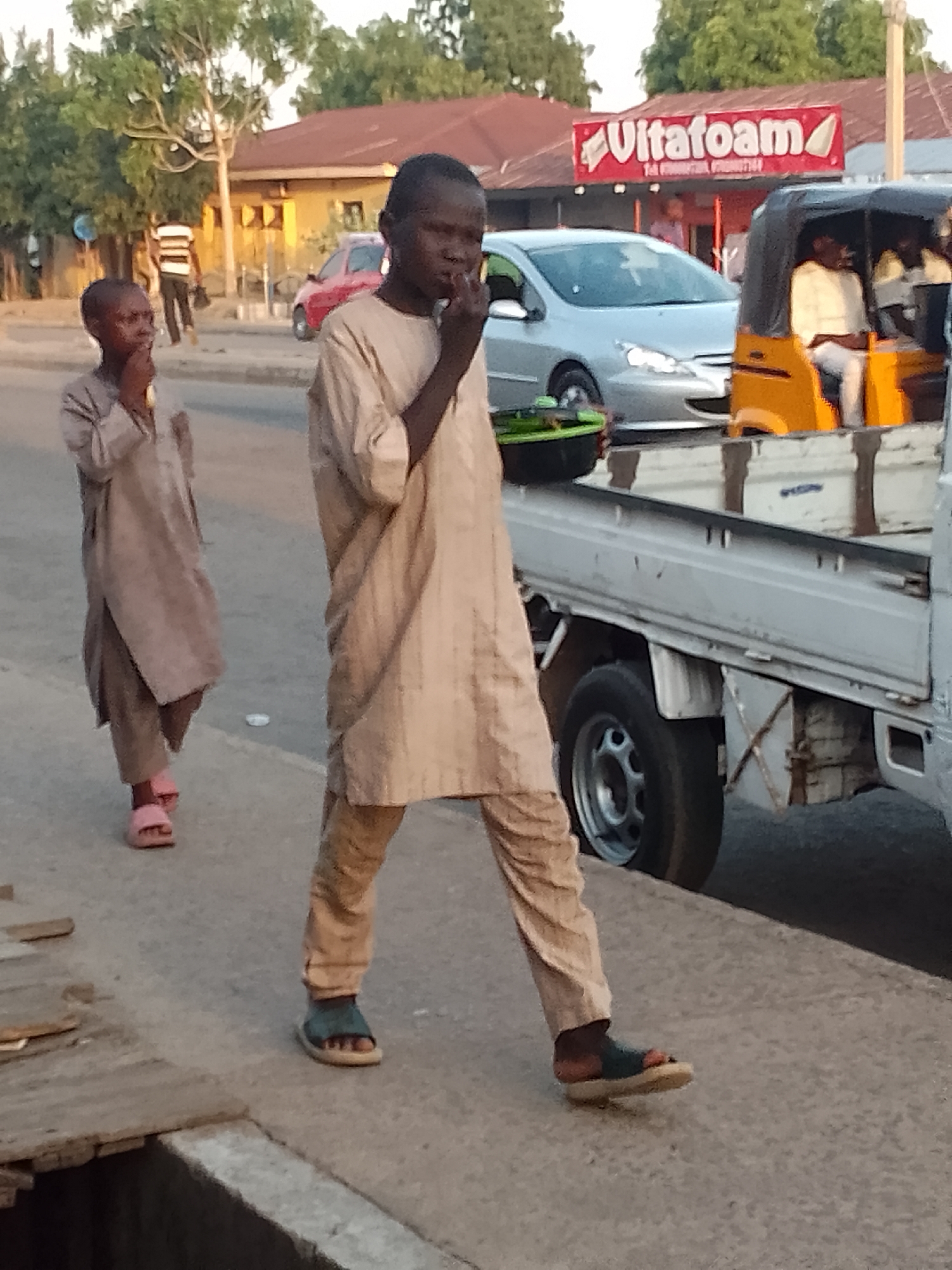 This image captures a street scene in Pantame, Nigeria, during daylight hours, likely late afternoon. In the foreground, two dark-skinned young males, approximately pre-teen age, walk from left to right on a dirt and concrete sidewalk. Both are dressed in light-tan, loose-fitting traditional attire consisting of trousers and long-sleeved tunics. The male closer to the camera, on the right, wears dark blue/green sandals and holds a small, possibly green, object or container in his left hand while his right hand touches his mouth/face. The male further back on the left wears pink slides and also touches his mouth/face with his right hand.
To their immediate left, a partially covered drain or open ditch with wooden planks is visible along the edge of the sidewalk. The paved road runs parallel to the sidewalk. On the road, a silver sedan, likely a Peugeot model, is visible in the mid-ground, facing left. Behind it, a yellow and black auto-rickshaw (Keke Napep) with at least two passengers is partially visible, with the bed and rear wheel of a white light-duty flatbed truck occupying the lower right foreground.
In the background, a prominent red sign with white lettering displaying "Ultrafoam" and a partial phone number ("Tel: 0803...") is mounted on a building. Several other single-story buildings with red or light-colored roofs are visible amidst green trees and foliage. A person in a striped shirt and dark pants walks further back on the sidewalk, away from the camera. The overall scene depicts an active urban or semi-urban environment.