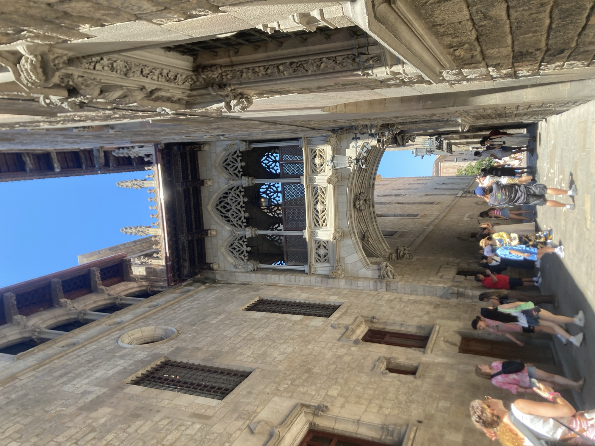 The image shows a view looking up a narrow street in Barcelona, Spain, towards a Gothic-style bridge connecting two buildings. The bridge has intricate stone carvings and open arches. The buildings on either side of the street are made of aged stone with arched windows and decorative architectural elements. A group of tourists are visible in the street below, some taking pictures and others walking. The sky above is clear and blue. The overall impression is one of historical architecture and a popular tourist destination.