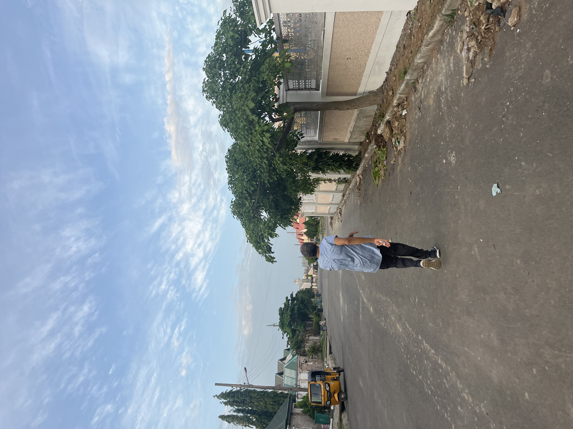 In Abuja, Nigeria, a person walks down a paved road in a residential area. The individual, wearing a blue collared shirt and dark pants, is viewed from behind, walking away from the camera. To the left of the road, a yellow three-wheeled vehicle is parked. Houses with green roofs line the street, interspersed with trees and decorative fences. The sky above is blue with scattered white clouds. The road is mostly clear of debris, though some scattered litter and construction materials are visible along the edges of the sidewalk. The overall scene suggests a quiet, suburban setting.