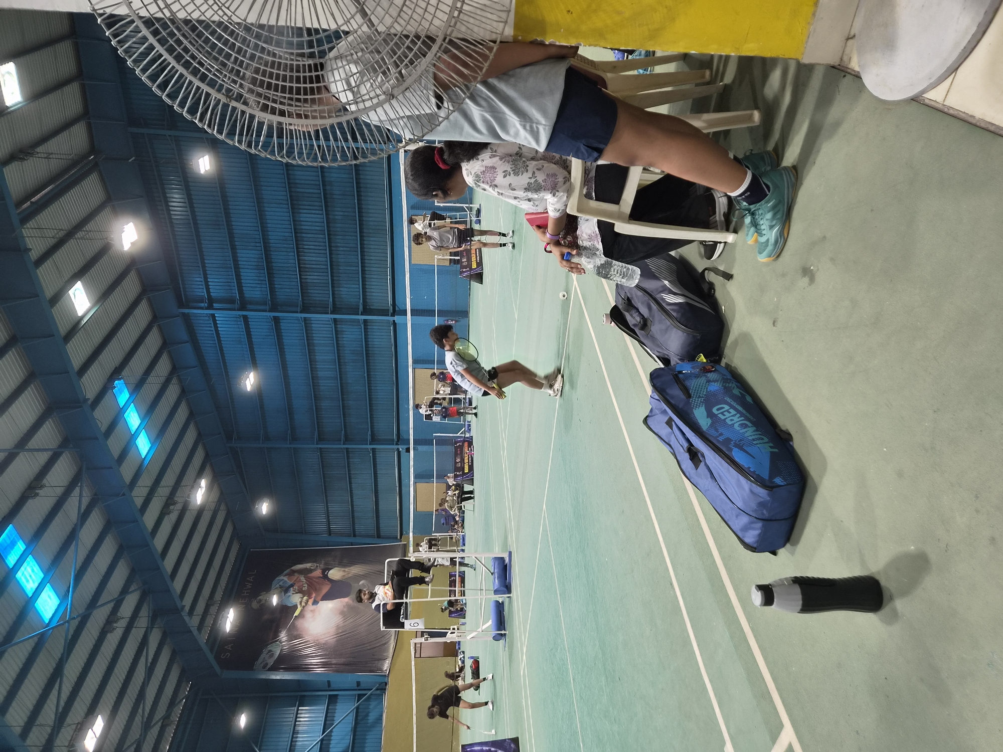 The image depicts an indoor badminton court in Phagwara, India. The court is illuminated by overhead lights, and the walls are lined with blue corrugated metal. A large banner featuring a badminton player is displayed on the back wall. Several individuals are present on and around the court. In the foreground, two large blue badminton bags and a water bottle are placed on the floor. To the right, a large industrial fan partially obscures the view of a woman sitting on a white chair, wearing a floral print top and dark shorts. Another person, wearing a light blue shirt and dark shorts, is also seated nearby, with their legs crossed. On the court, several badminton players are engaged in matches, including a player in a black t-shirt and shorts on the left, and a player in a grey t-shirt and shorts in the center. An umpire is seated on a raised chair, observing the game. Additional spectators are visible in the background. The court surface is a green synthetic material with white boundary lines.