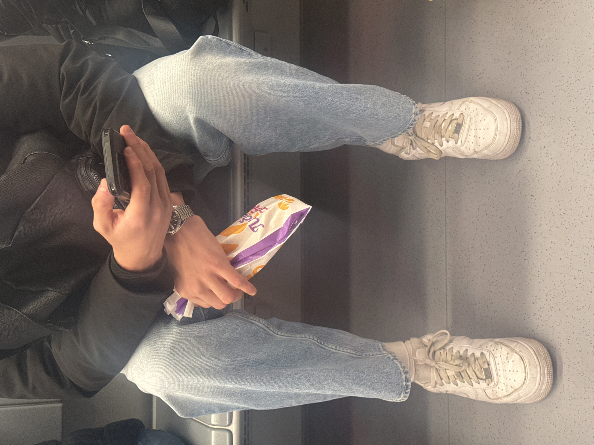 A person sits in an interior space, likely on public transport based on the seat visible in the lower left. The person wears light wash jeans, white sneakers, and a dark jacket. They are holding a cell phone in their left hand and a fast food item, identifiable as Taco Bell, in their right hand. A wristwatch is visible on their left wrist. The floor is a light grey with a speckled pattern. The location cannot be determined as the surroundings are not visible.