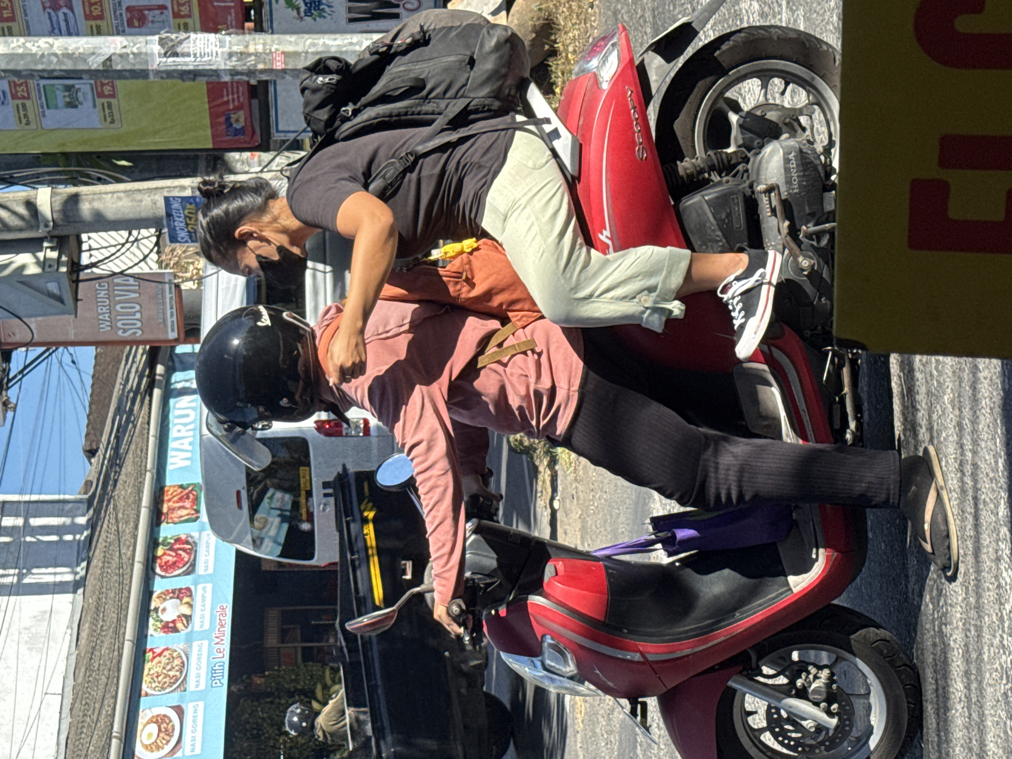 In Kuta Selatan, Indonesia, a woman with dark hair pulled back and wearing a black face mask rides on the back of a red scooter. She is dressed in a dark grey t-shirt and light green pants. She is holding onto the shoulders of the scooter's driver, who is wearing a pink long-sleeved shirt, black pants, and a black helmet. A large black backpack is on the woman's back. The scooter is in motion on a paved street, with a black truck and a white van visible in the background. A banner above the truck reads "WARUNG" with images of food. Other signs with Indonesian text and pricing are visible on utility poles to the right. The scene appears to be a typical street view in a bustling Indonesian town.
