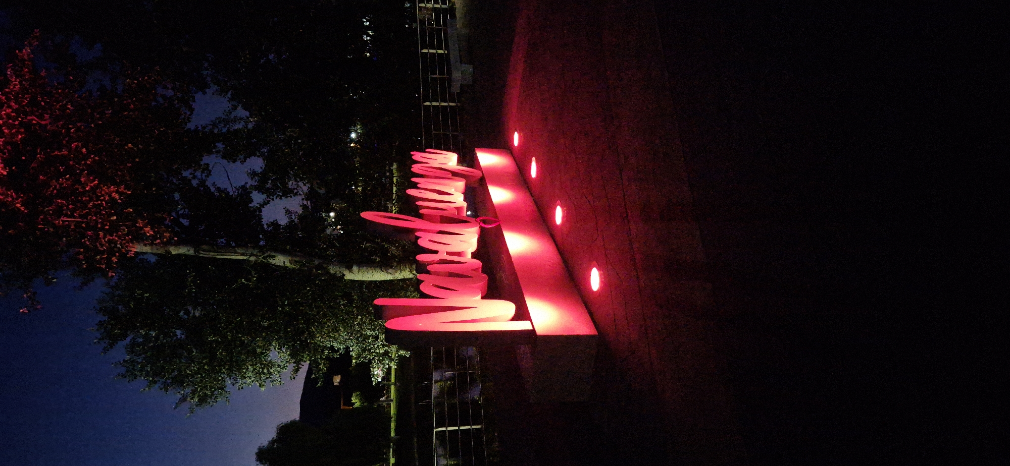 The image shows a neon sign in the city of Navaluenga, Spain, at night. The sign is illuminated in bright pink and red lights and is positioned on a dark-colored base. Several circular lights are embedded in the ground in front of the sign, also emitting a red glow. In the background, trees with dark foliage are visible, with some branches illuminated by a red light. The sky above is a deep blue, indicating it is nighttime. A body of water with a railing is also faintly visible in the mid-ground, suggesting the sign is located near a river or lake. The overall scene is dark, with the sign and ground lights being the primary sources of illumination.