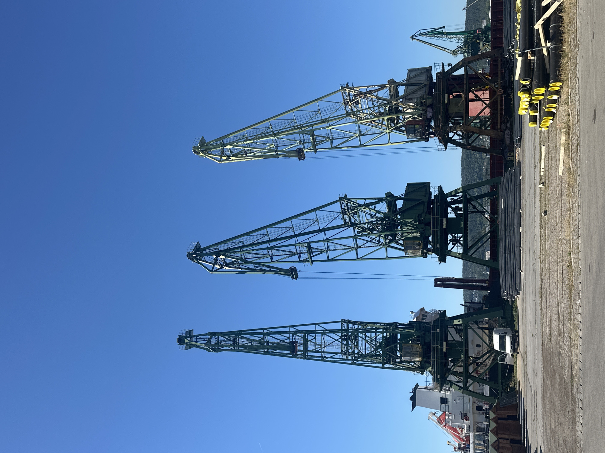 The image shows a port scene under a clear blue sky. In the foreground, there are stacks of black pipes with yellow caps. A paved area with tire tracks leads towards a row of small wooden structures on the left and a white van parked in front of large industrial cranes. Three large green industrial cranes dominate the mid-ground, with their lattice structures reaching high into the sky. Behind the cranes, a large white ship with a red stripe is docked. In the background, a green hillside is visible with some buildings. A smaller green crane can be seen on the far right, further in the distance. The scene appears to be a busy industrial port.