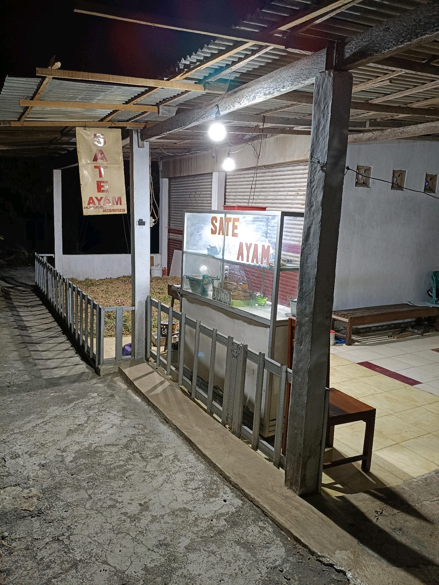 This image shows an outdoor food stall at night, likely in Semanu, Indonesia, based on the text on the banner. The stall is under a corrugated metal roof supported by wooden beams and concrete pillars. A banner hanging from the roof reads "SATE AYAM" (Chicken Satay) in large red letters, with a phone number below it. In front of the stall is a glass display case with what appears to be food items inside. A metal fence encloses the seating area, and a wooden bench and small table are visible in the background. The ground is concrete and somewhat uneven. The lighting is provided by bare bulbs hanging from the ceiling.