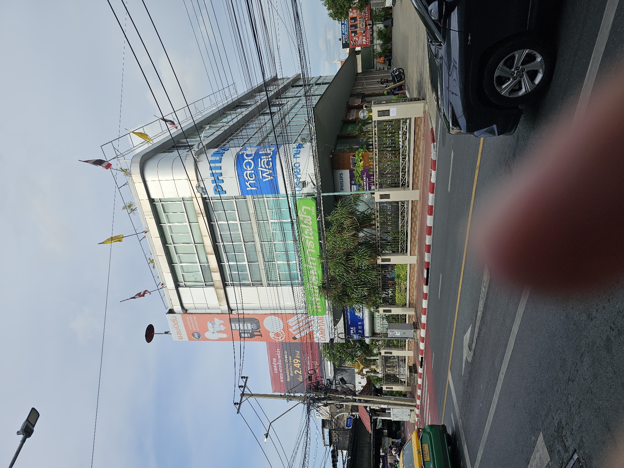 The image depicts a street scene in Bangkok, Thailand, with a prominent multi-story building featuring large signs for "PHILIPS" and other Thai text. The building is modern with a glass facade. Several flags, including the Thai flag and yellow flags, are visible on the rooftop. Overhead, a dense network of electrical wires crisscrosses the sky.
In the foreground, a dark-colored SUV is parked on the right side of the road. A yellow taxi is visible on the left side, moving away from the camera. The road has lane markings and a pedestrian crossing with red and white stripes.
Various smaller signs and advertisements are present on the building's lower levels and on poles along the street. The environment appears to be a commercial or business district, with lush green trees planted along the sidewalk in front of the building. The sky is overcast.