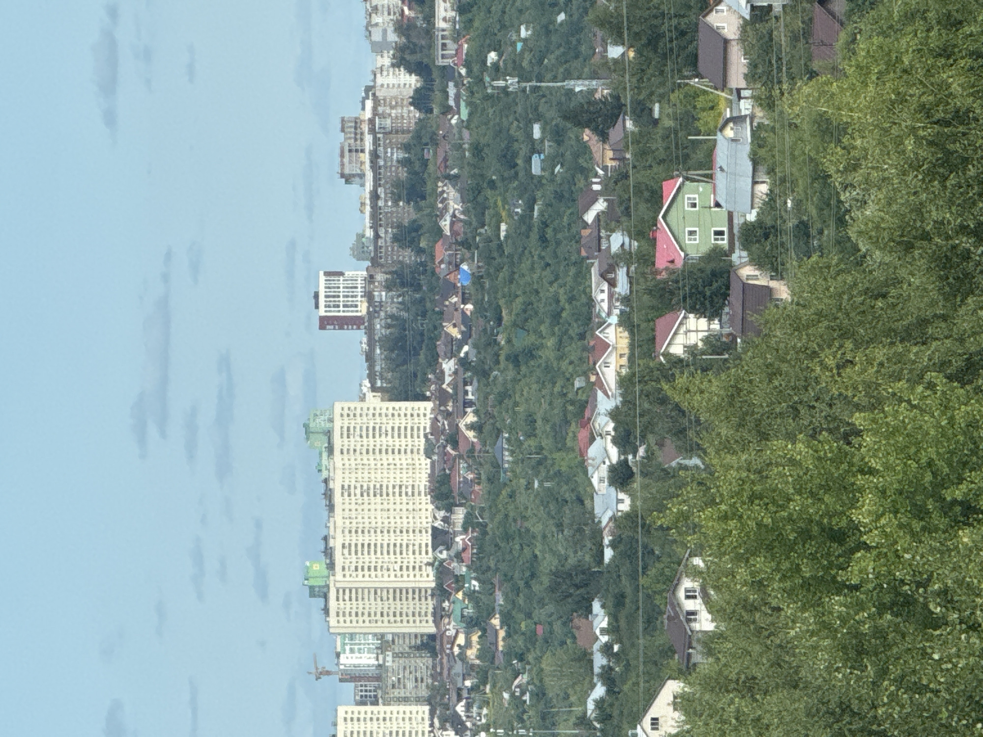 This is a medium-range, eye-level, vertical, outdoor photograph taken on an overcast day, depicting a dense suburban or exurban residential area with varying architectural styles. In the background, several multi-story apartment buildings are visible, some with light-colored facades and others with darker, more modern designs. One prominent building is tall and light beige with a grid-like pattern of windows. Another building features a white and red striped facade.
In the middle ground, a densely wooded hillside is covered with numerous residential houses, many of which are relatively small and appear to be detached single-family homes. The houses display a variety of roof colors, including red, gray, and brown, and their architectural designs range from traditional to more contemporary. Some houses have visible windows and chimneys. The overall impression is one of a sprawling development interspersed with significant greenery.
The foreground is dominated by lush green trees, likely deciduous, with full foliage, obscuring some of the lower parts of the houses. Power lines are visible crisscrossing the scene horizontally in the mid-ground and foreground, indicating the presence of electrical infrastructure. A telecommunications tower is also present in the middle ground, to the right of center. The sky is a pale blue with scattered, wispy clouds. No people are visible in the frame. The scene suggests a residential landscape in or near Анкудиновка, Russia, characterized by a mix of apartment complexes and a proliferation of detached homes amidst natural vegetation.
